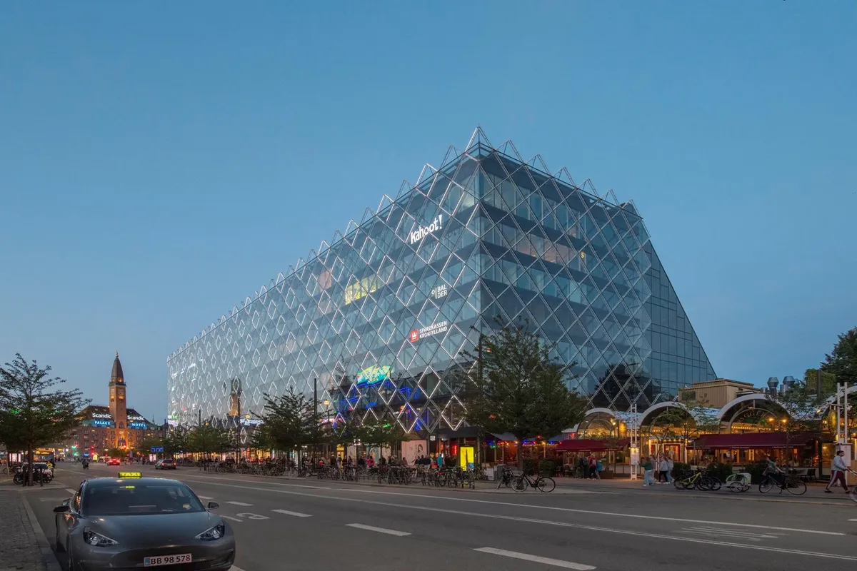 En bil kører langs en gågade med restauranter og et stort, glasklædt bygning med teksten "Kahoot" på facaden
