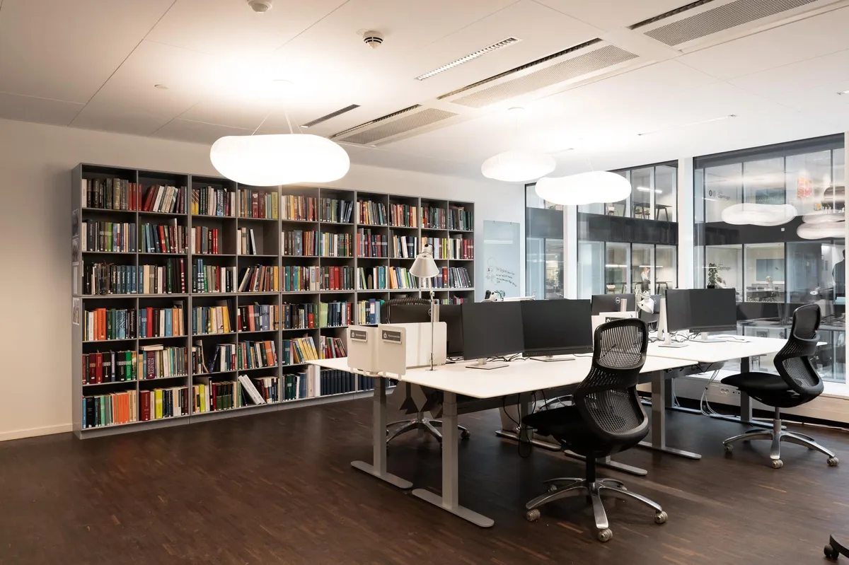 Kontorrum med lyse loftslamper og en stor boghylde med mange bøger og flere computere med skærme