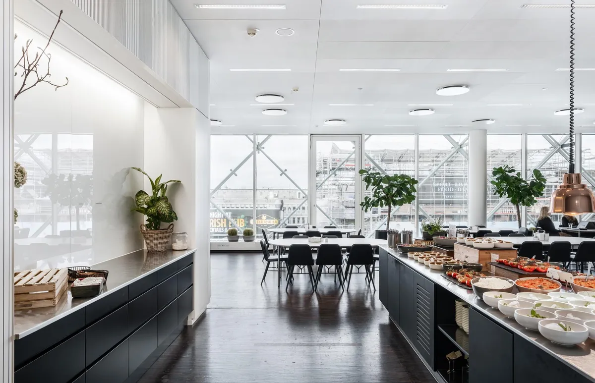 Kontorlokaler med moderne indretning, plante og træhylder, lyse lofter, bord og stole samt spisebord med mad