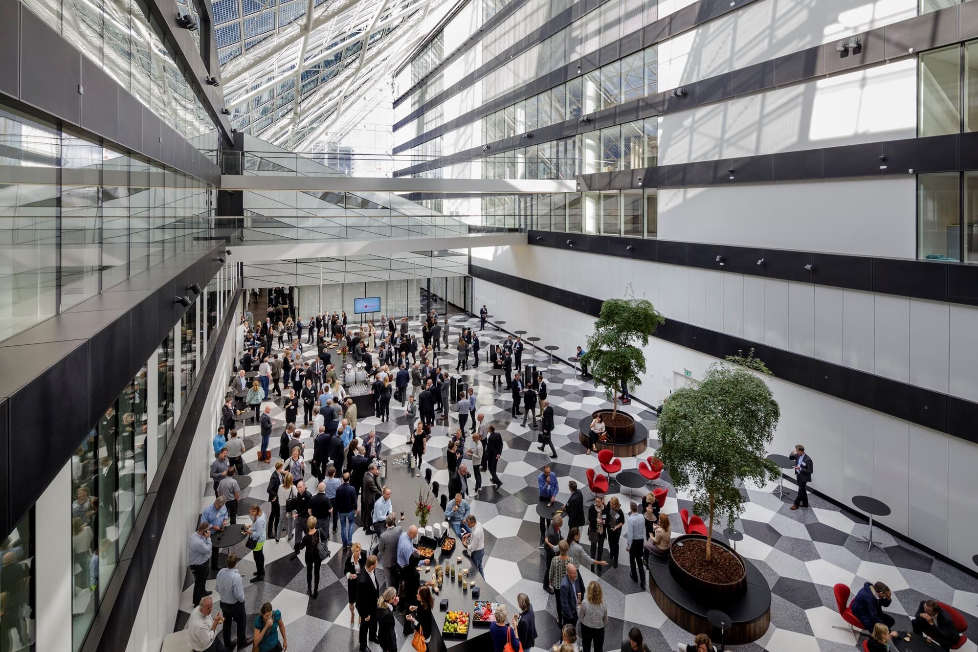Stor lobby med grupper af folk, der samles, diskuterer og nyder mad og drikke, forskellige plantetyper og møbler placeret i