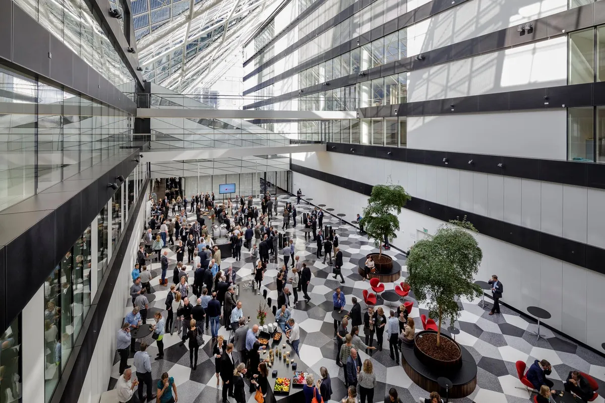 Stor lobby med grupper af folk, der samles, diskuterer og nyder mad og drikke, forskellige plantetyper og møbler placeret i