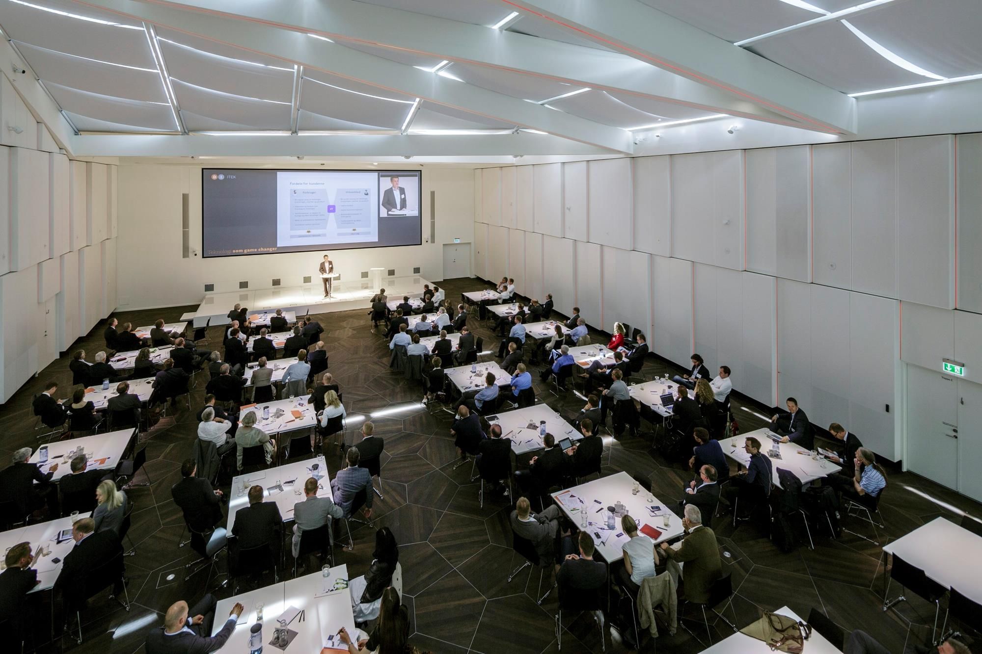 Konferencerum med højt til loftet og moderne møbler, hvor en mand holder foredrag for en gruppe tilhørere