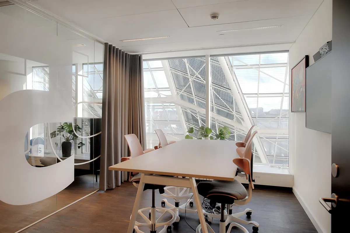 Kontorlokaler med lyse glasvægge og trægulv, moderne møbler og udendørs udsigt gennem vinduer og solfanger