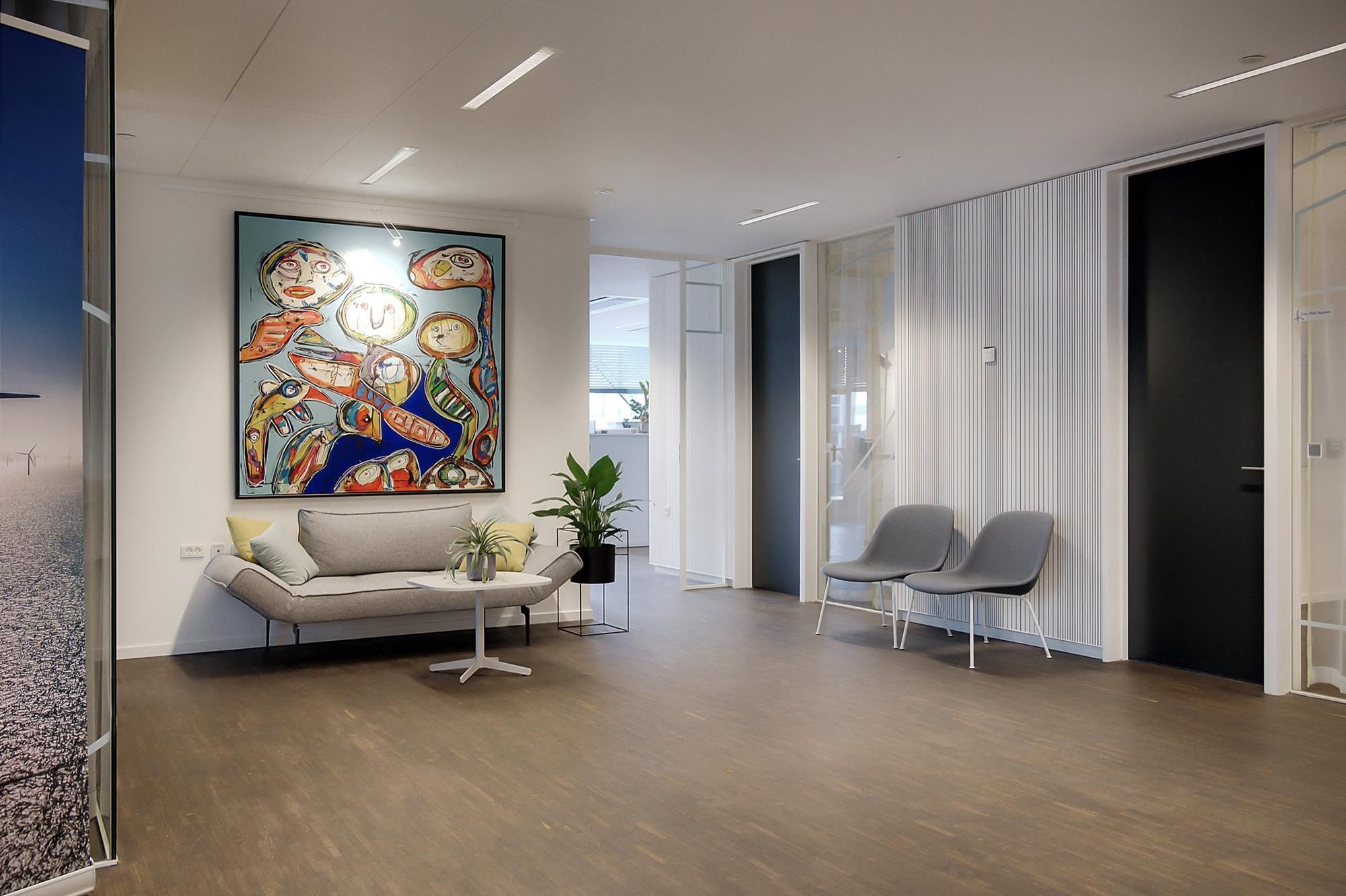 Kontorlokaler med lyse vægge, hvor der står en sovesofa med pude og en bord med planter og en lampe samt en stor maleri.