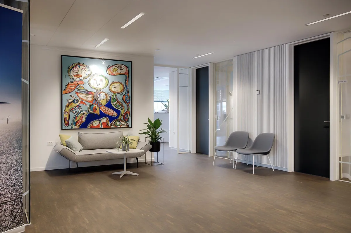 Kontorlokaler med lyse vægge, hvor der står en sovesofa med pude og en bord med planter og en lampe samt en stor maleri.
