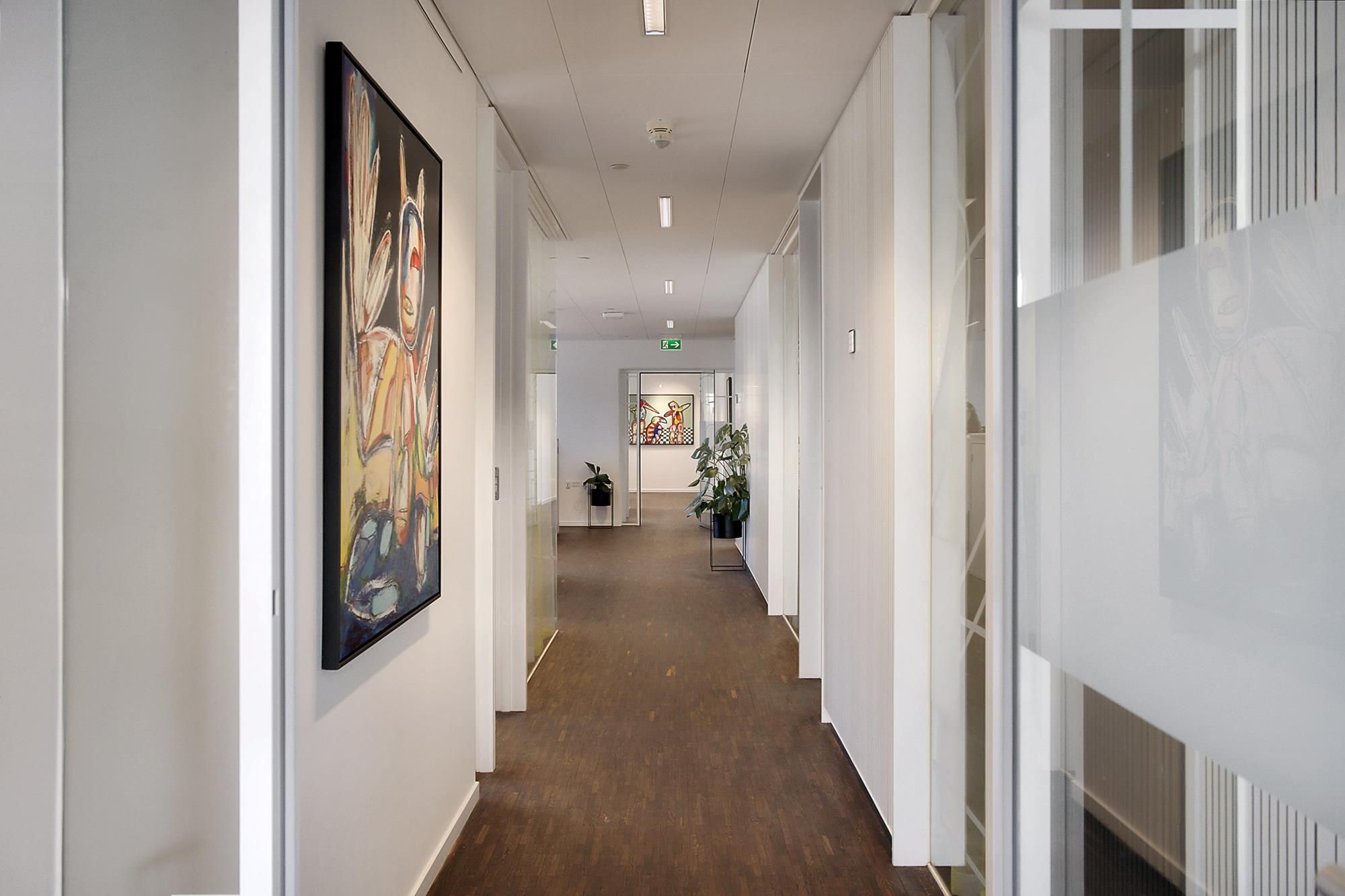 Kort og lyst kontor med lyse lofter og kunst på væggen samt planter og glasdøre