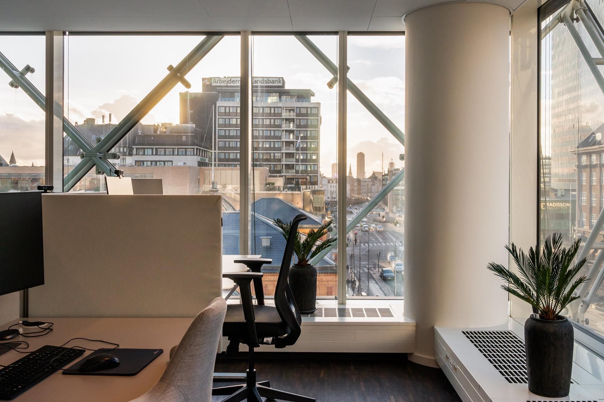 Kontorrum med udsigt til byen, skrivebord med computer og plante, glasvinduer og højtliggende bygninger i baggrunden