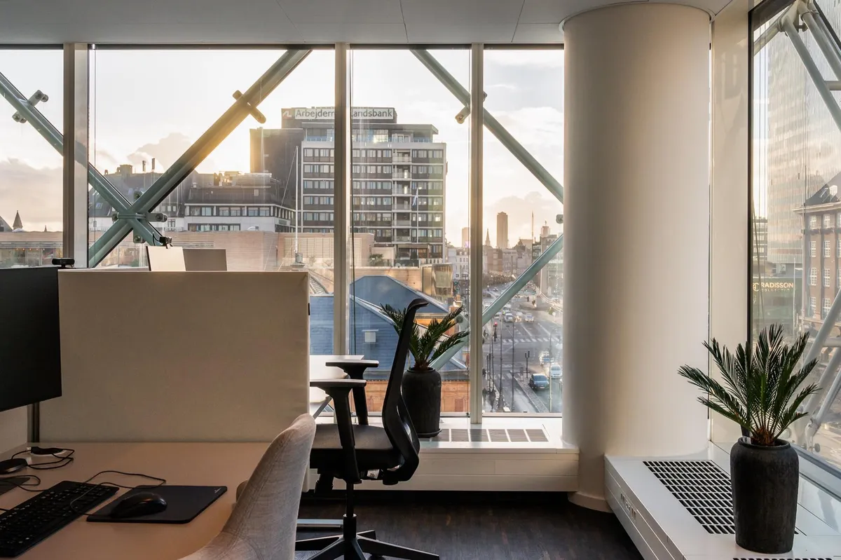 Kontorrum med udsigt til byen, skrivebord med computer og plante, glasvinduer og højtliggende bygninger i baggrunden