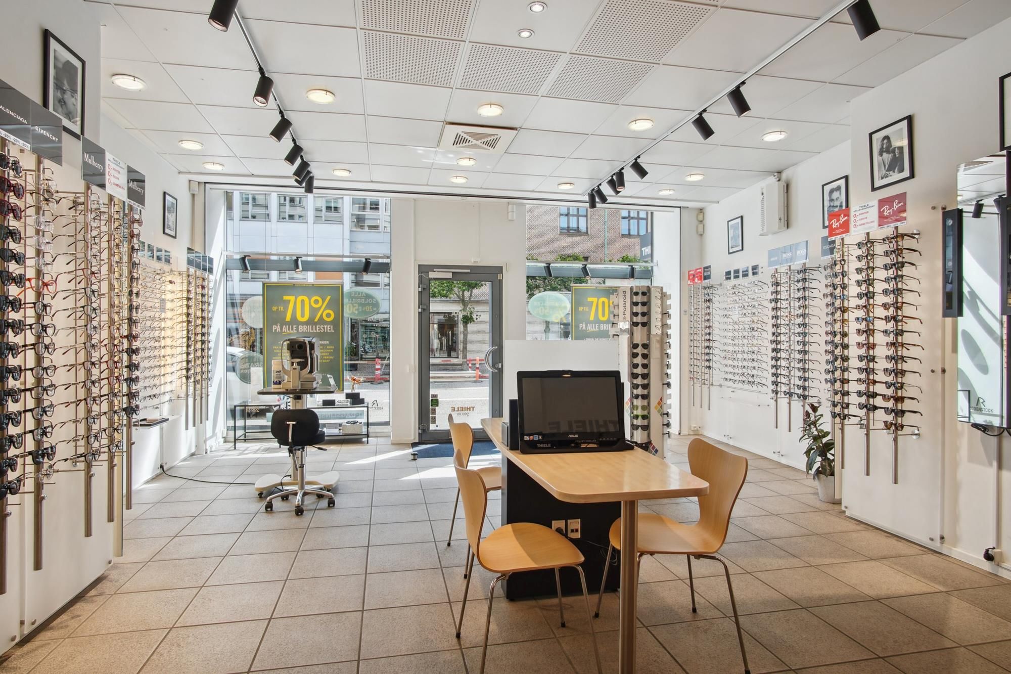 Udenfor en butik er der en række solbriller og briller monteret på væggen, og der er en monitor og et bord med stole indenfor