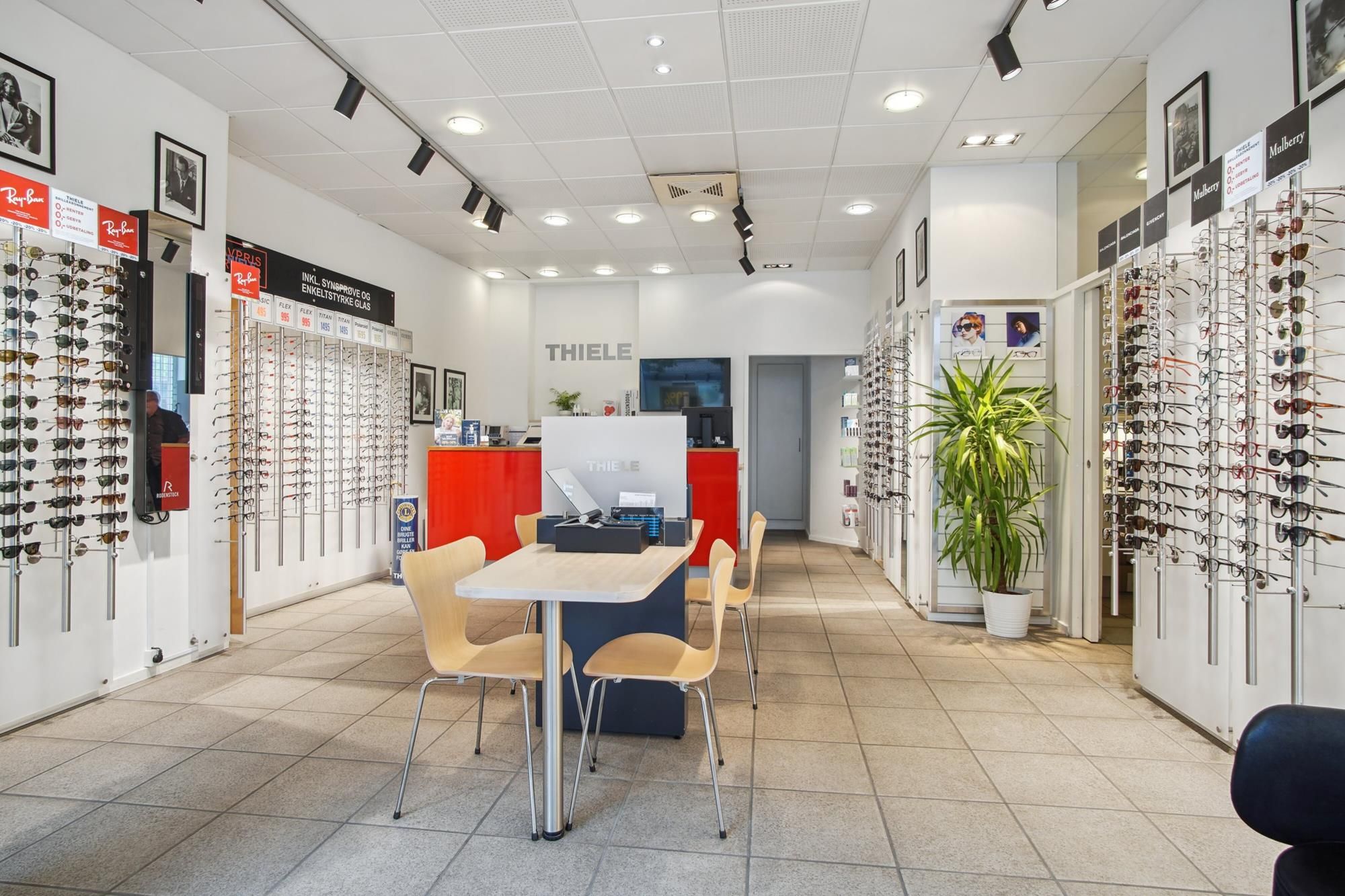 En moderne brillebutik med briller i skuffer og et lille salgsbord med stole og plante i hjørnet