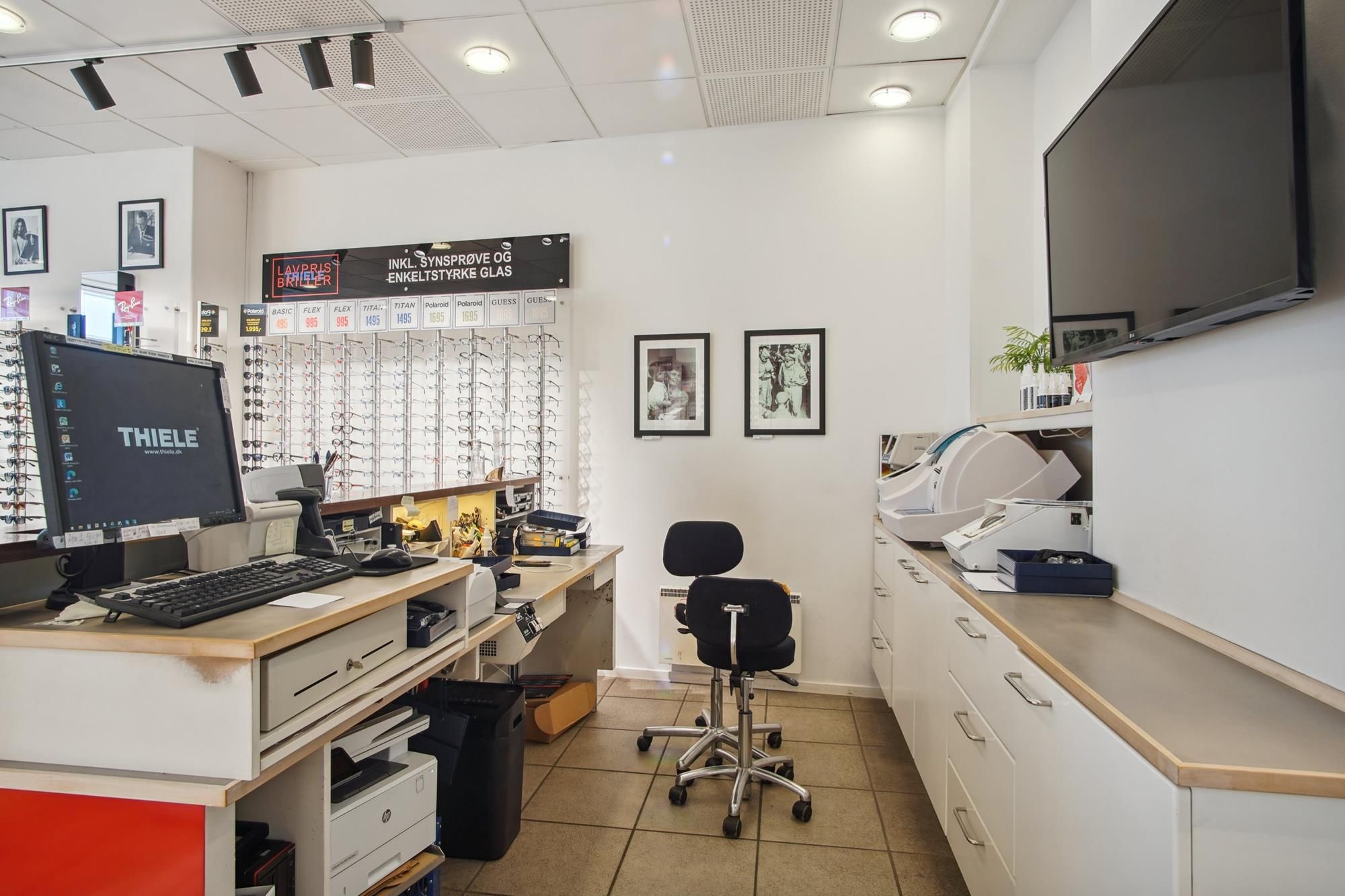 Kontor med to skriveborde, hvor et har en computer, og et stol er udstillet med briller på væggen og et fjernsyn