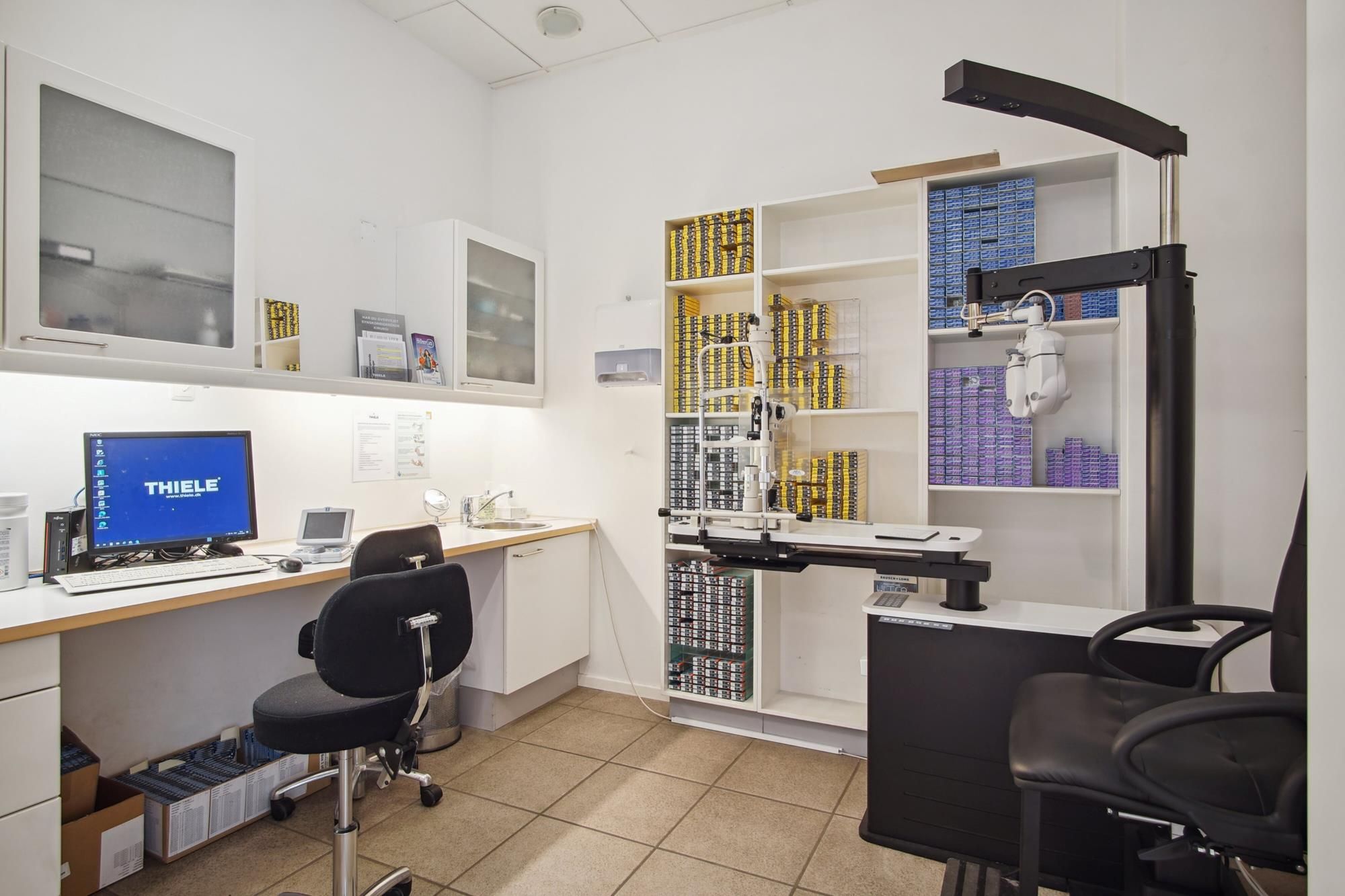 Kontor med moderne computer og skrivebord, to stole og en høj hylde med optikudstyr og briller