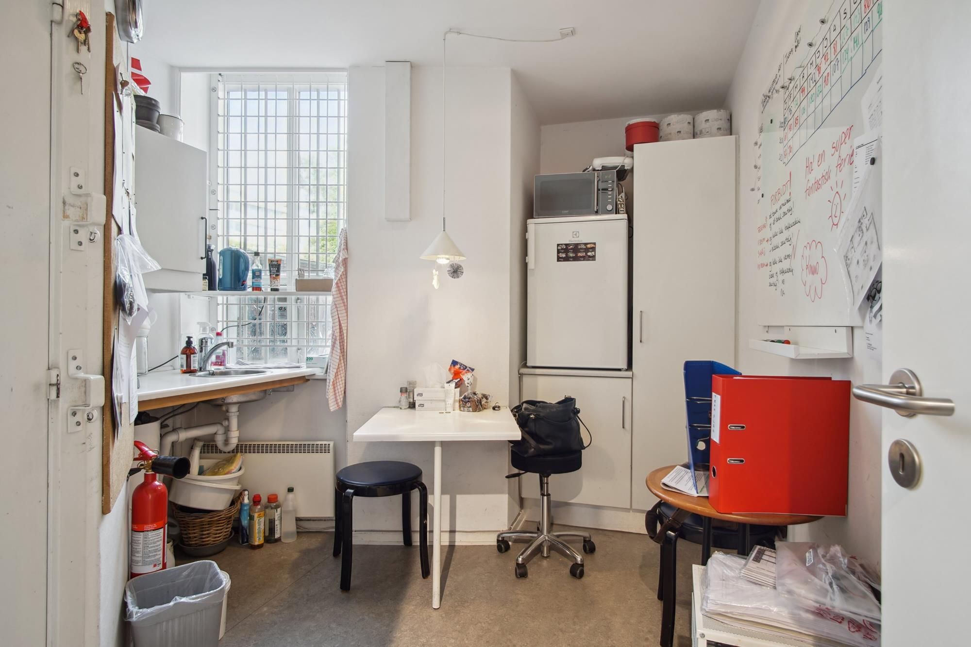 Et lille rum med et hvidt bord, en taske og en højstol, en kalender og en hvid køleskab og en ildslukke