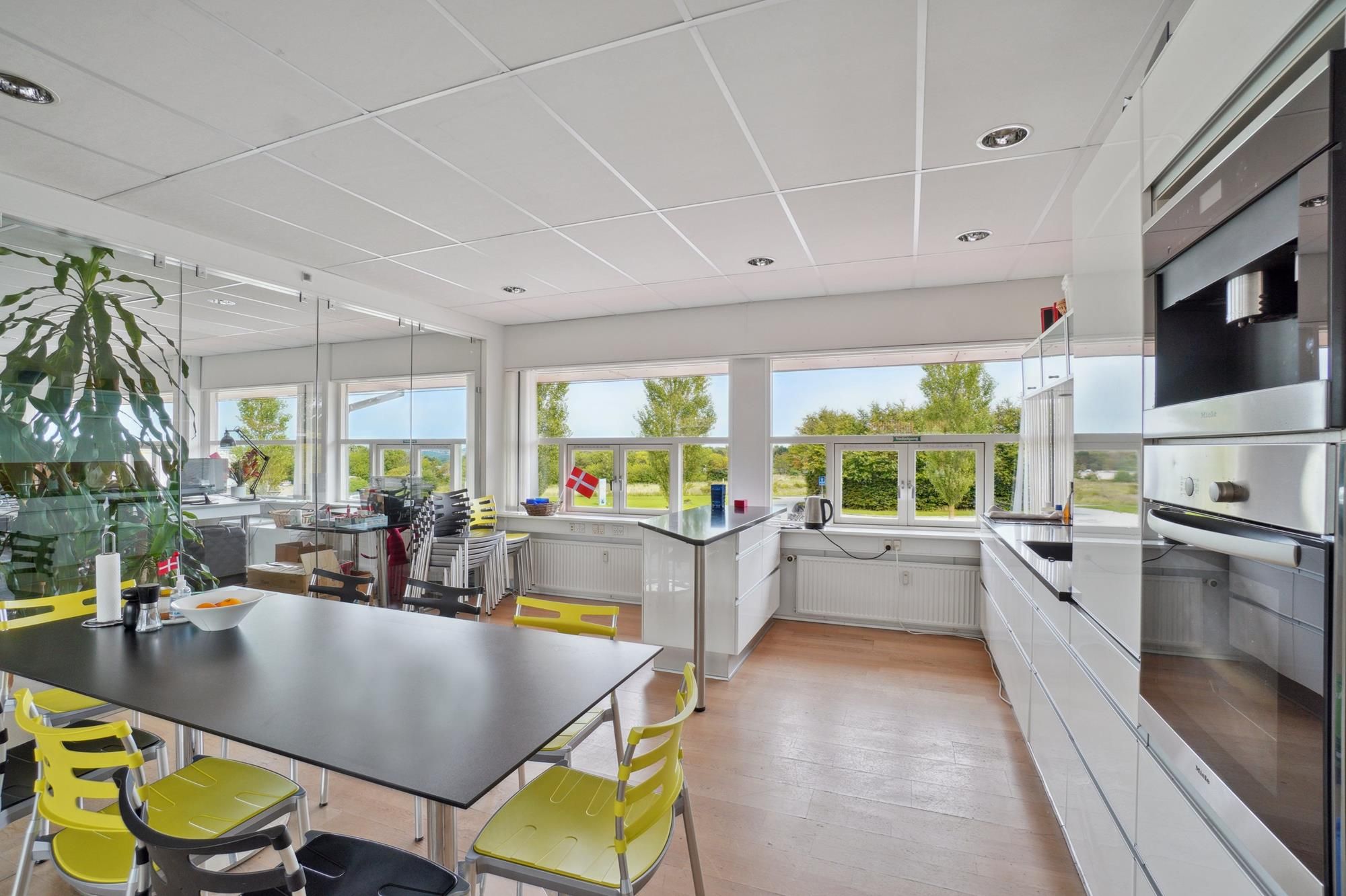 Moderne køkken med højt loft, stort vindue og stolpe med flag og lysegul stol og lysegul bord med frugtskål og servietter