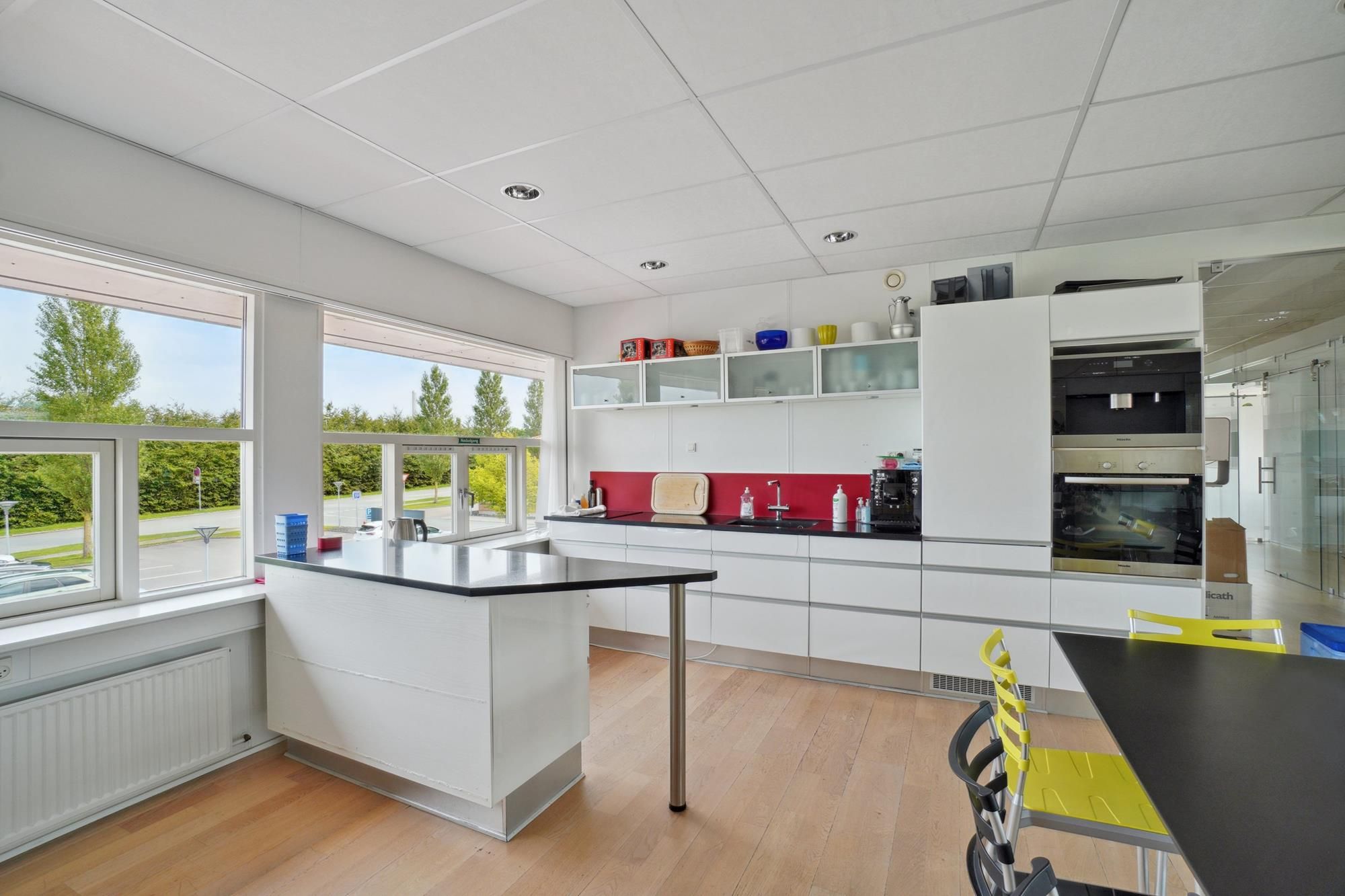 Moderne køkken med højt loft, hvide vægge, gult gulv og store vinduer med udsigt til grønne træer og veje