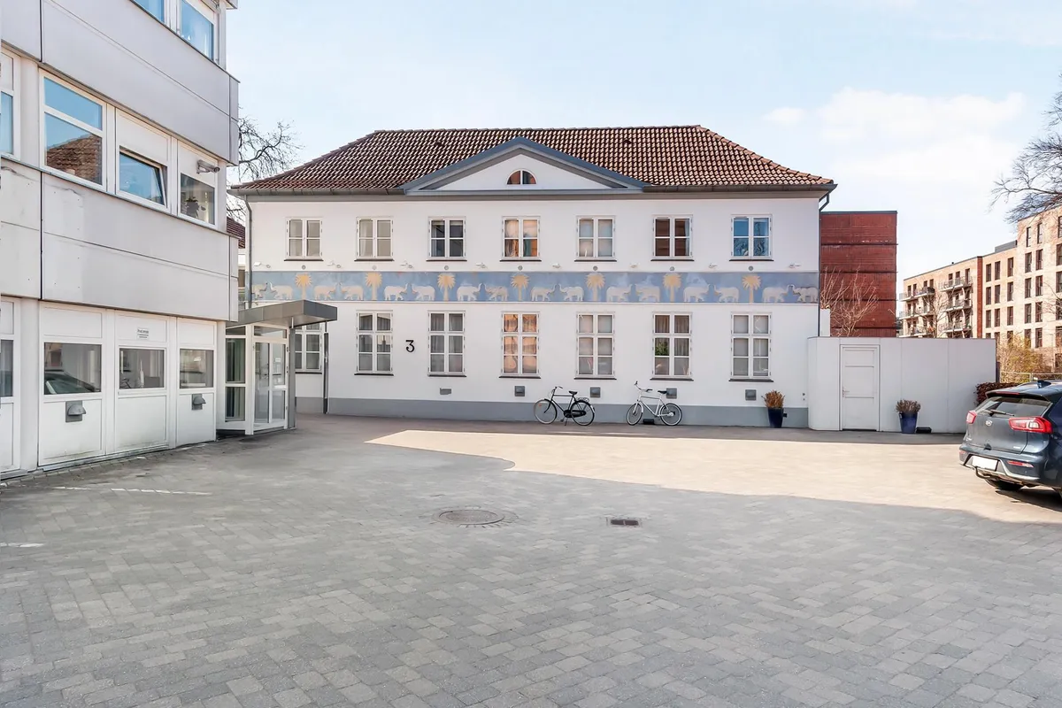 Moderne hvidt bygningsværk med glasruder og terrasse. Parkeringsplads med to cykler og bil. Søjle og buske i forgrunden.