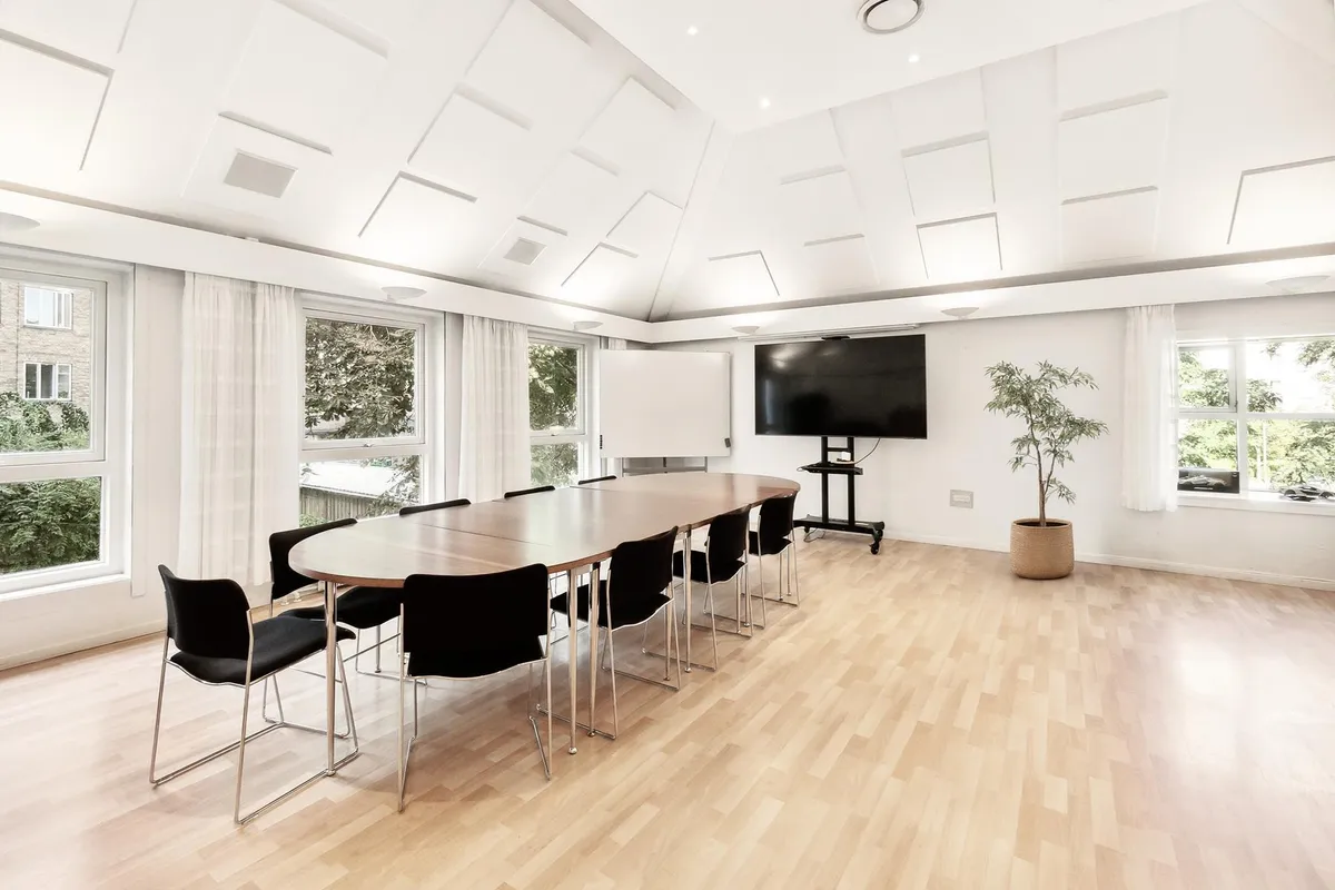 Luksuskontor med ovalt bord, 12 stole, tv, plante, hvide vægge, vinduer med hvide gardiner og træer udenfor