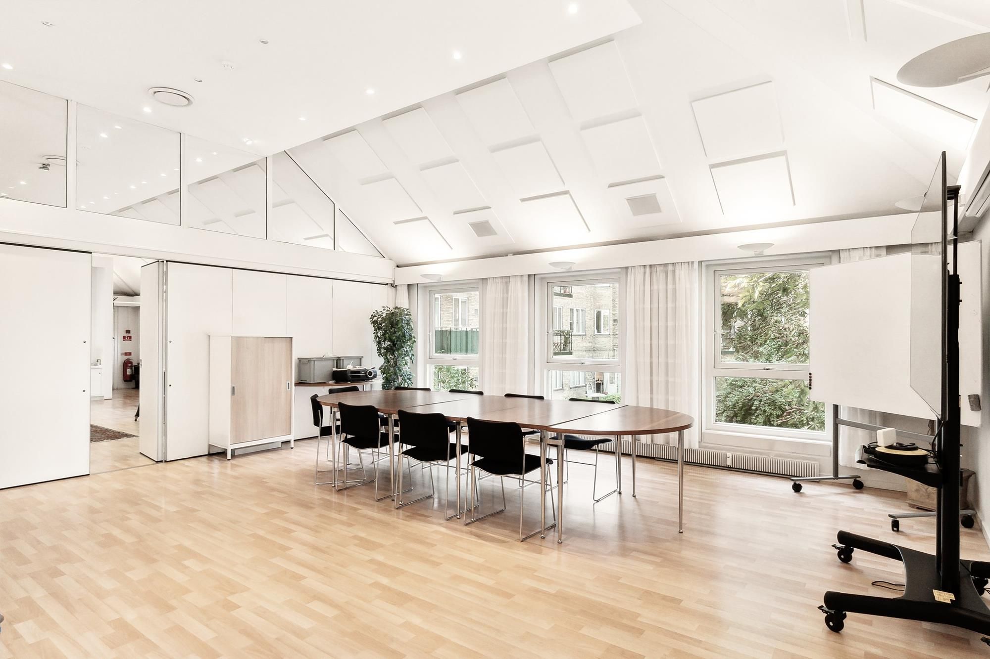 Et moderne mødelokale med hvide vægge og trægulv. Rundt om er der flere lyse, hvide loftsreflekser. Væggen er dekoreret med