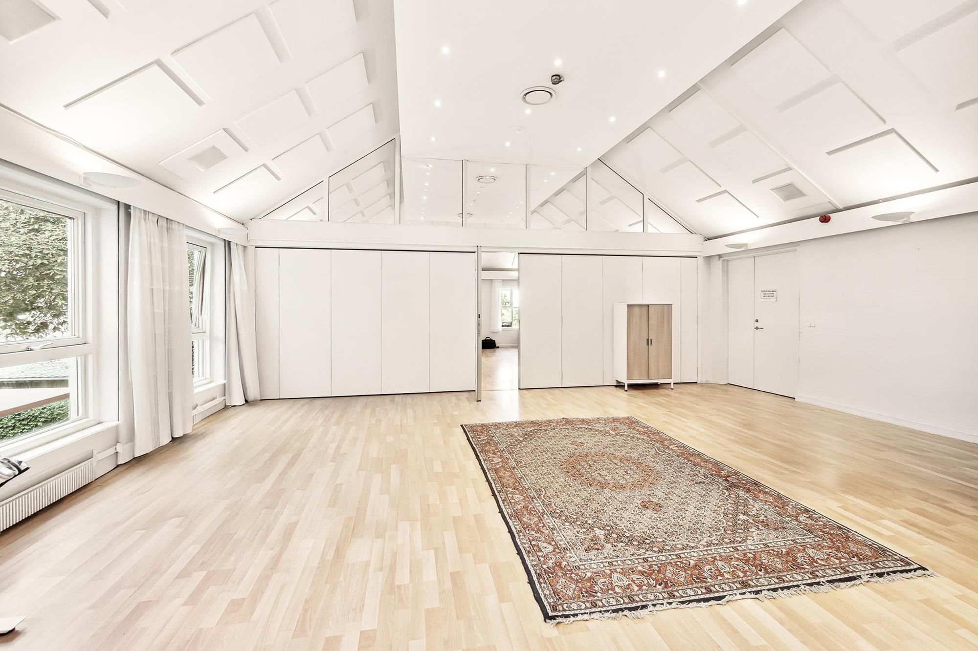 En tomt kontor med hvide vægge og trægulv, et orientalsk tæppe og store glasvinduer med hvide gardiner