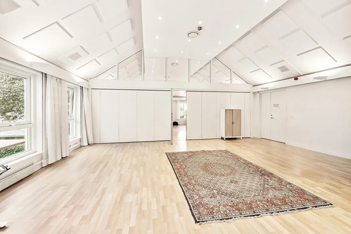 En tomt kontor med hvide vægge og trægulv, et orientalsk tæppe og store glasvinduer med hvide gardiner