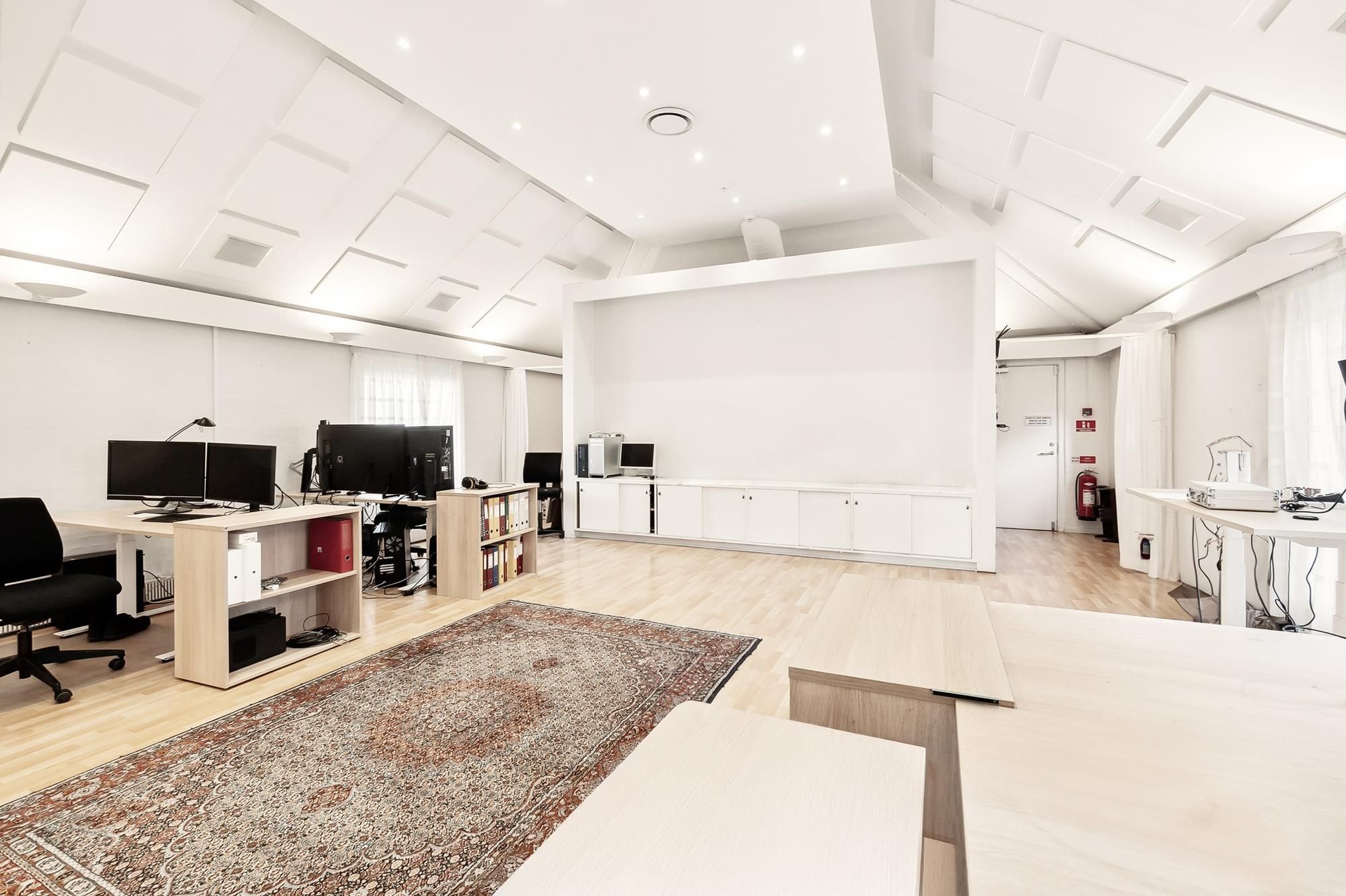 Luksuskontor med lyse vægge og trægulve, møbler og to skærme, højttalere og en orientalsk tæppe på gulvet