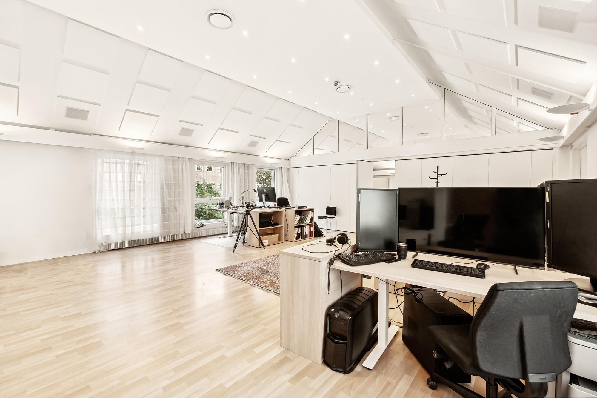 Kontor med flere skriveborde og skærme. Et skrivebord med to skærme og en computer. Kontor med højloft og lyse hvide vægge.