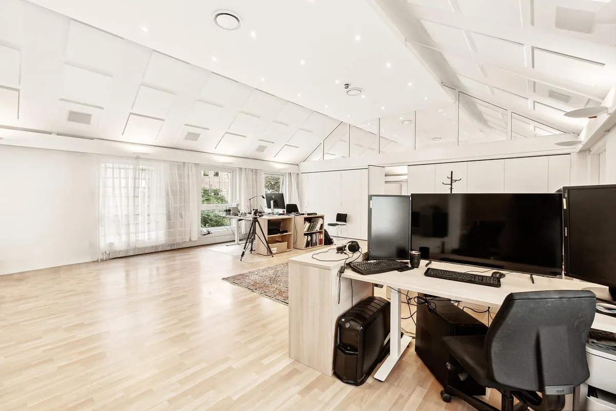 Kontor med flere skriveborde og skærme. Et skrivebord med to skærme og en computer. Kontor med højloft og lyse hvide vægge.