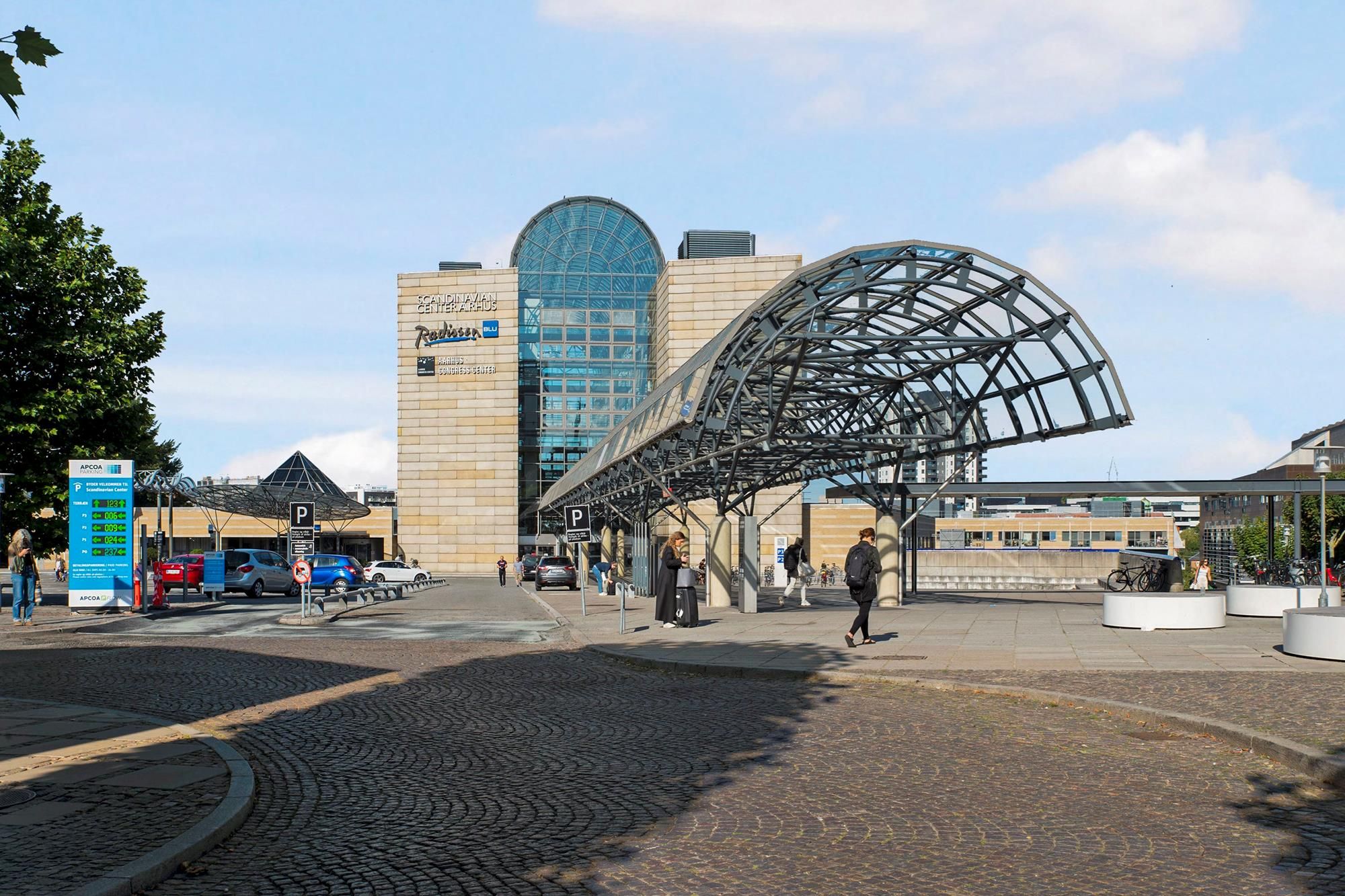 Bygning med moderne facade, glas og metalstrukturer -Mennesker går og står, nogle med tasker -Parkeret biler og schild med