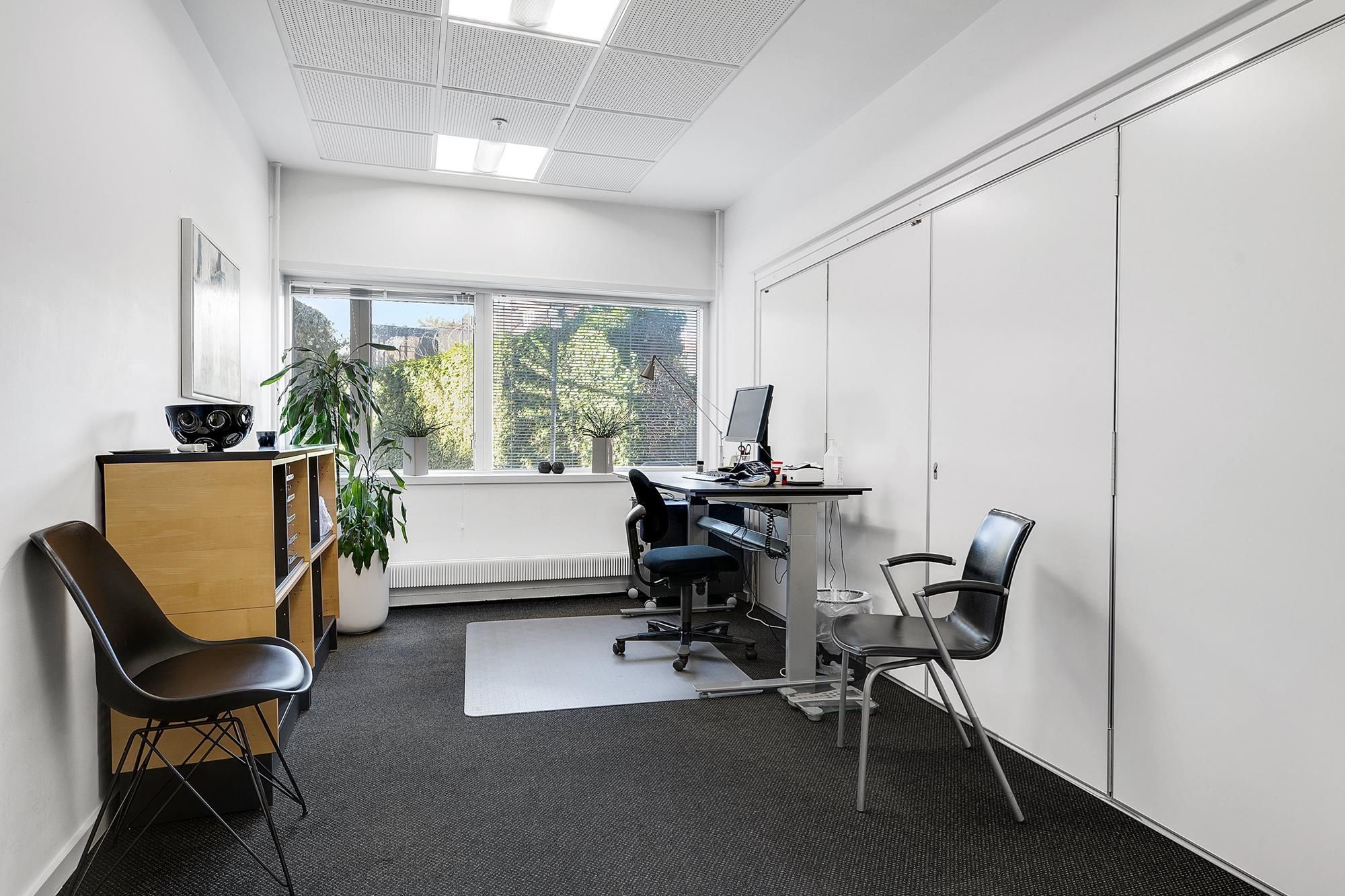 To moderne kontorstolar i sort og en høj højdeskriver med en computer og en lampe står i et hvidt kontorlokale med glasruder