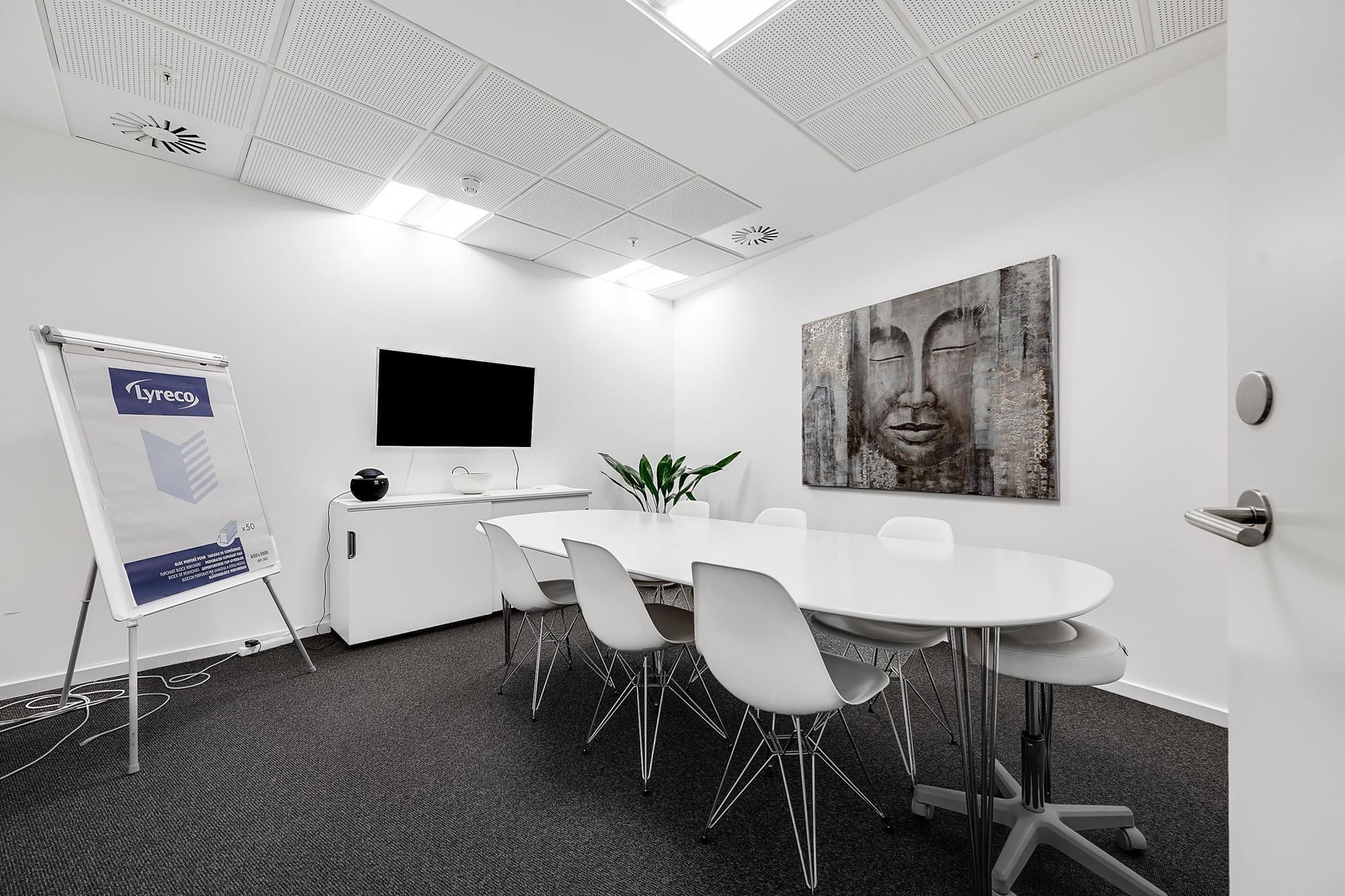 Mødeværelse med hvide vægge, hvide stole og rundt bord samt en skrivestand og tv-skærm på væggen