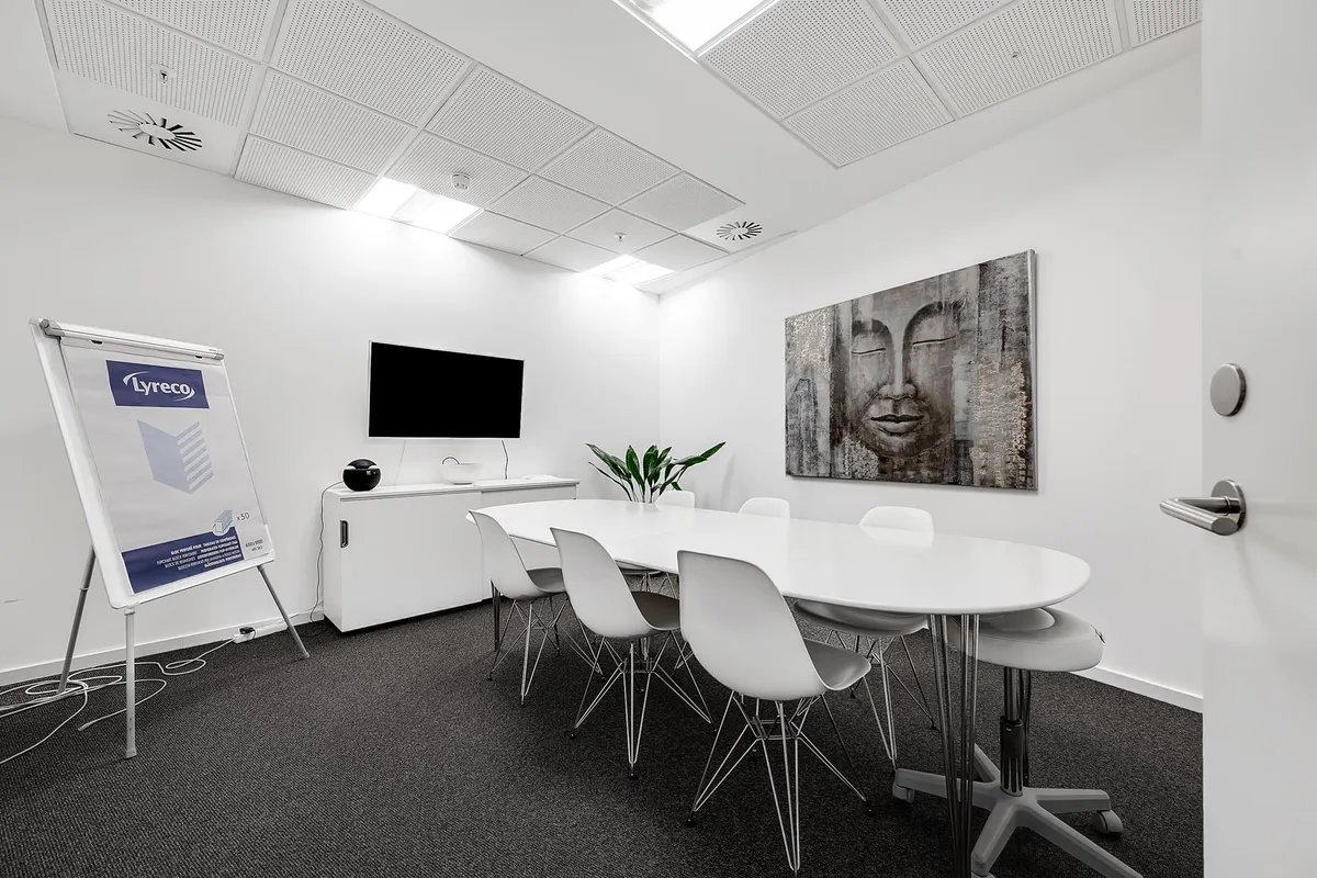 Mødeværelse med hvide vægge, hvide stole og rundt bord samt en skrivestand og tv-skærm på væggen