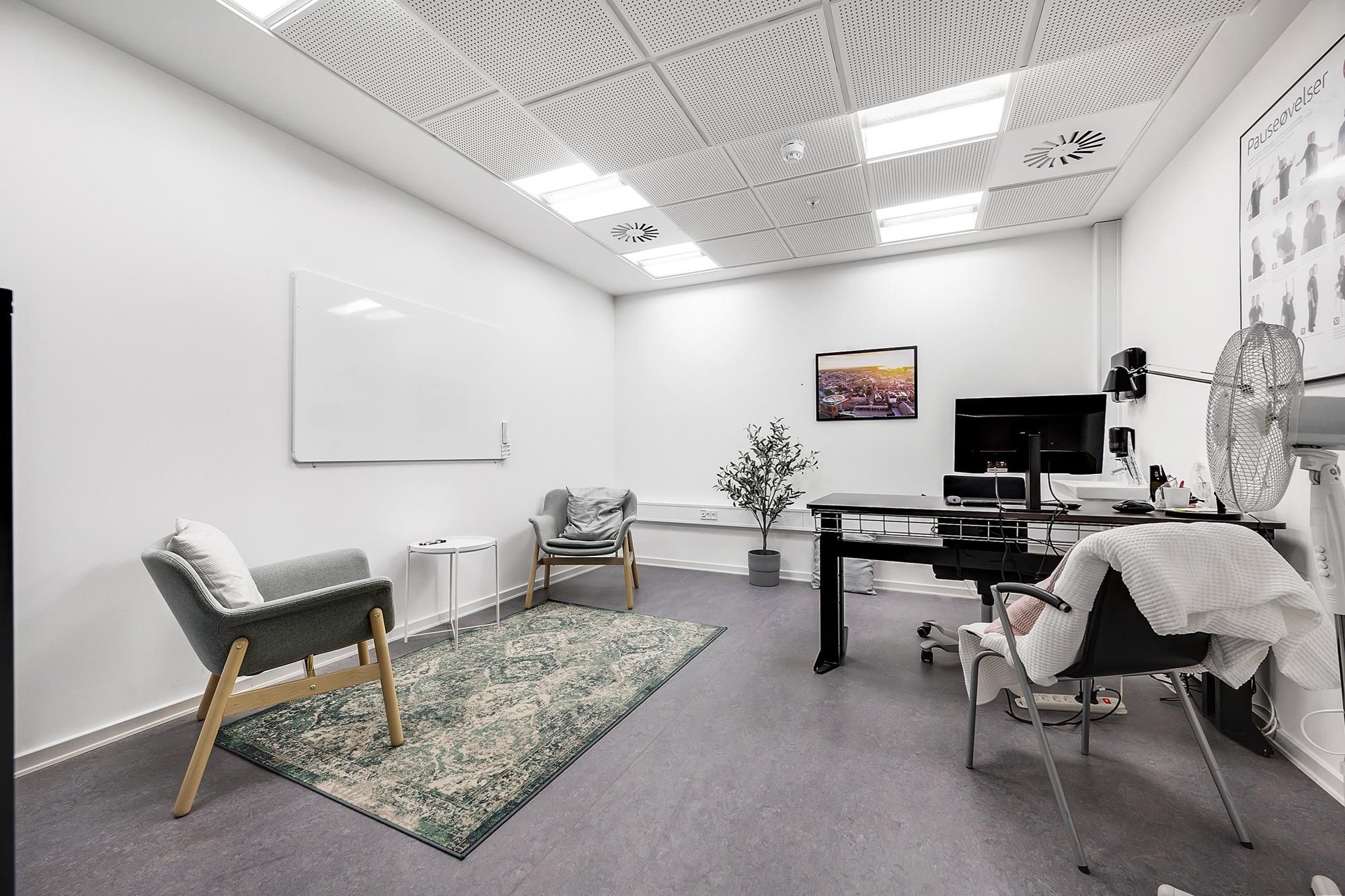 En enkelt klinik med en skrivebord og to stole med pude, hvor der er en lille plante og et billede på væggen