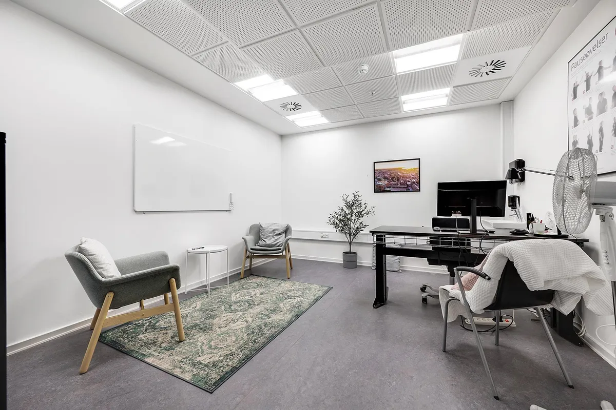 En enkelt klinik med en skrivebord og to stole med pude, hvor der er en lille plante og et billede på væggen