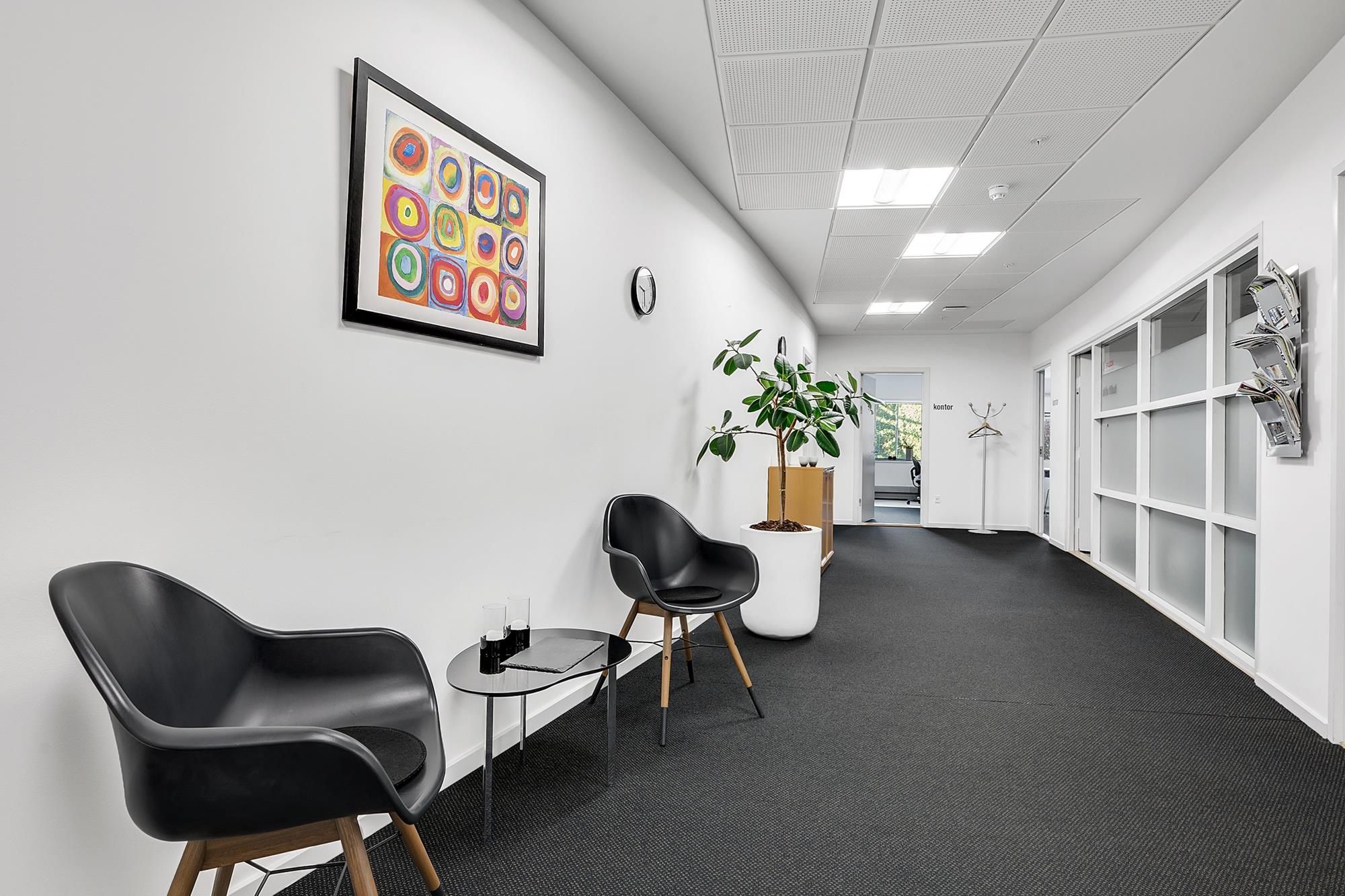 Moderne og velholdt klinik med hvide vægge og sortgule billeder, lænestol, plante og åben dør til et nyt rum med tøjstativ