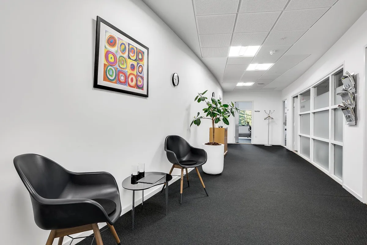 Moderne og velholdt klinik med hvide vægge og sortgule billeder, lænestol, plante og åben dør til et nyt rum med tøjstativ
