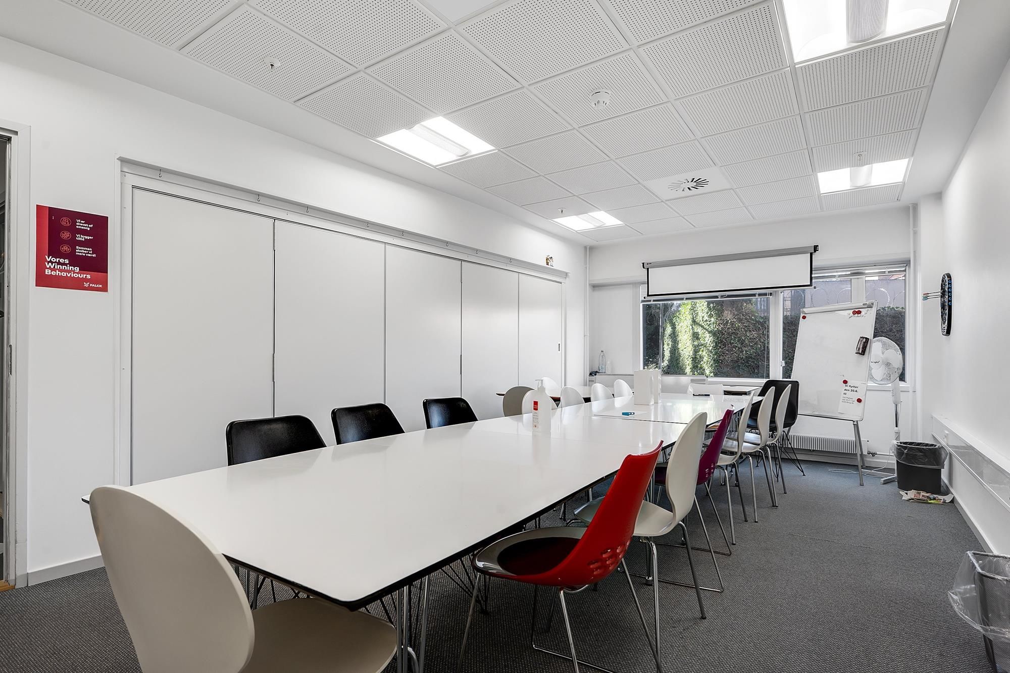 En stor og velholdt møde- og konferencelokale i klinikken med lyse vægge og hvide møbler samt skrivebord og radiatorer langs