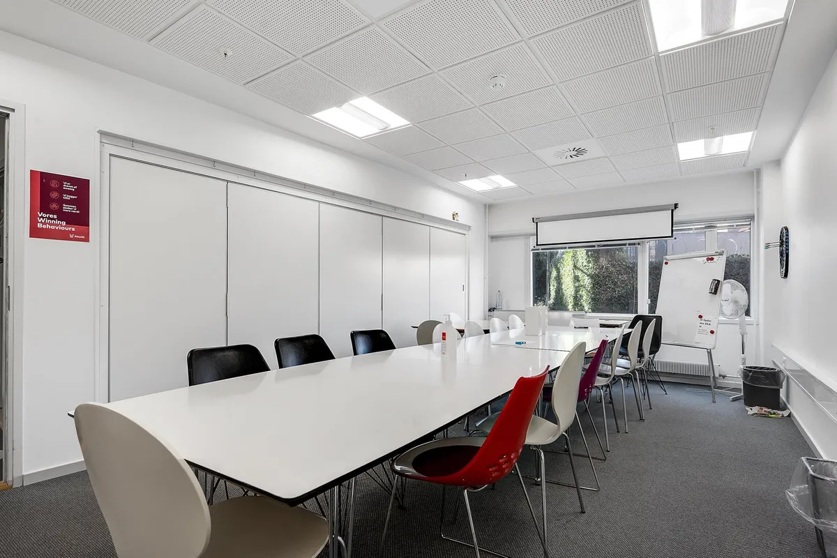 En stor og velholdt møde- og konferencelokale i klinikken med lyse vægge og hvide møbler samt skrivebord og radiatorer langs