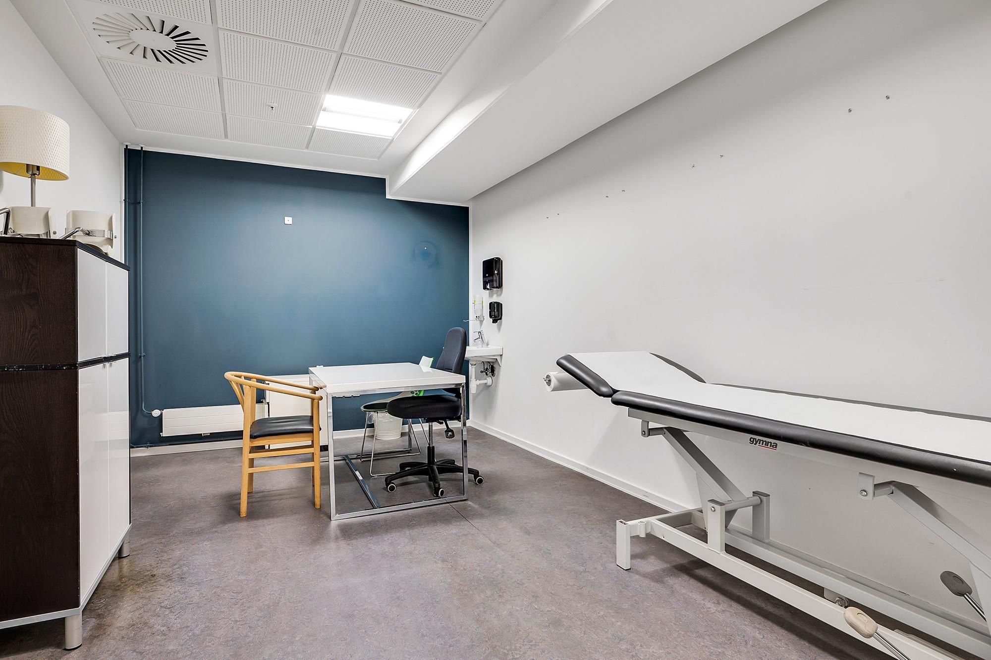 Ledig og velholdt klinik med hvid og blå væg, lægebord og gynastbord med håndtag, træstole og en lampe i hjørnet, hvor en