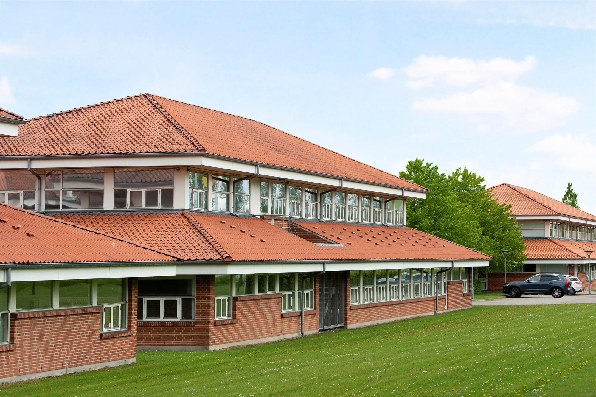 Lejlighedsbygning med rødt tegltag og glasvinduer står i en grøn park med parkerede biler i baggrunden