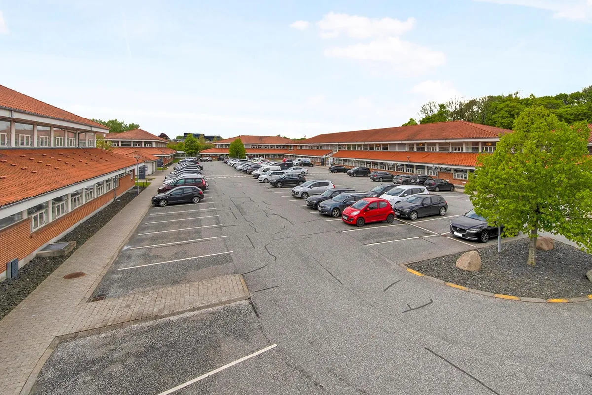 Parkeringsplads med mange biler foran en bygning med orange tag og glasvinduer