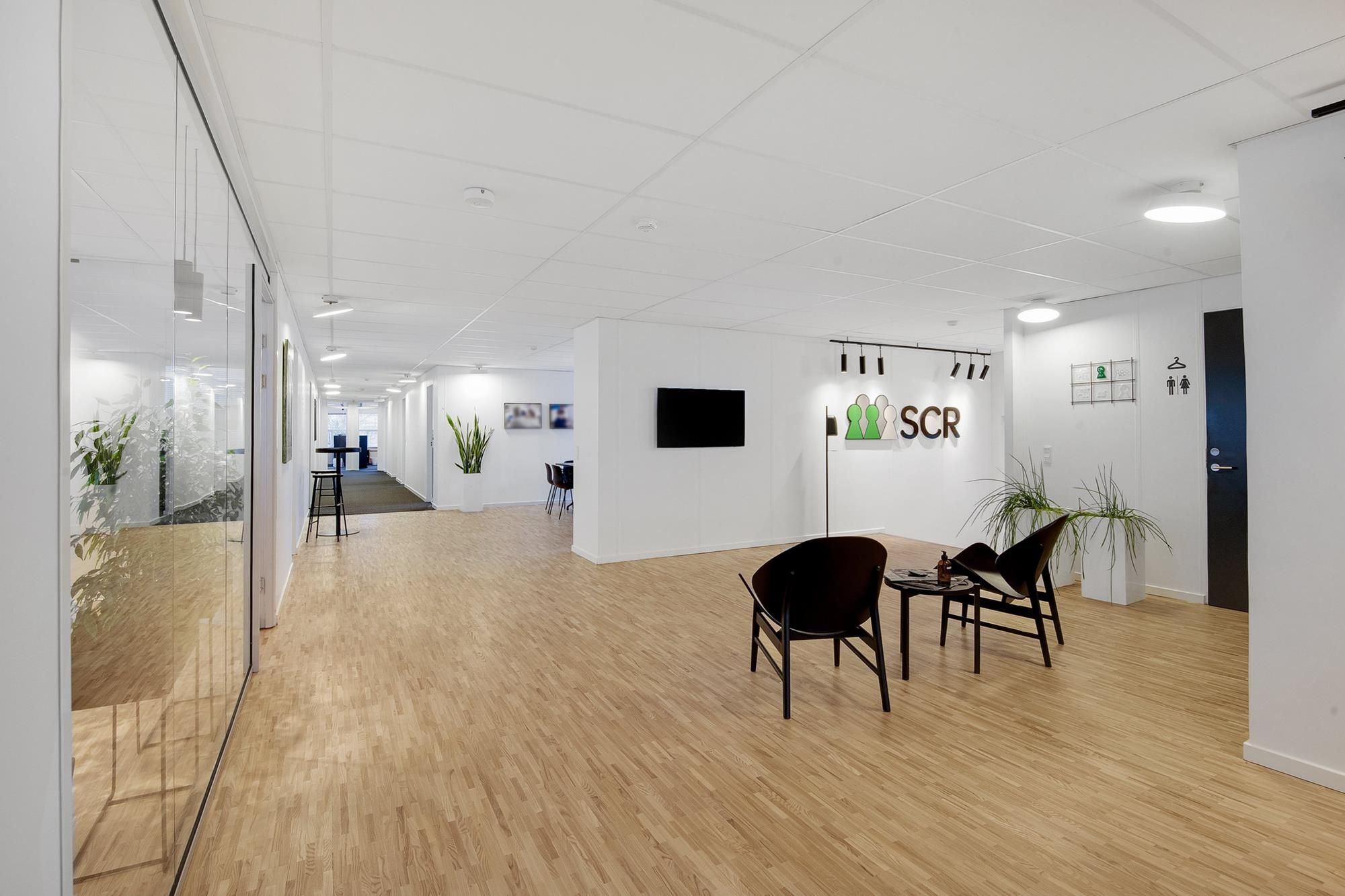 Moderne kontorlokaler med højloftede rum og hårdt trægulv, lyse vægge, og en stor vinduesfront med masser af naturligt lys