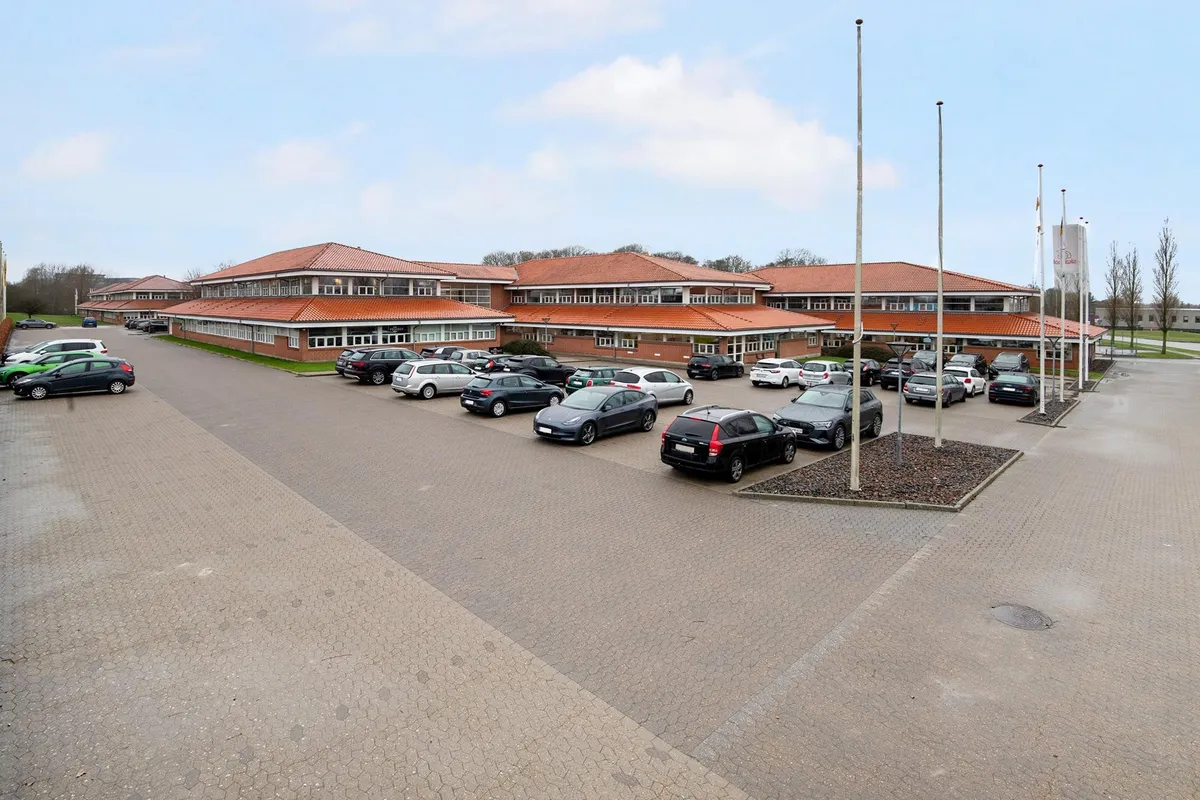 En stor bygning med røde tag er omgivet af parkeringspladser og biler, hvor flere flag er rejst og nogle bøsser står i en