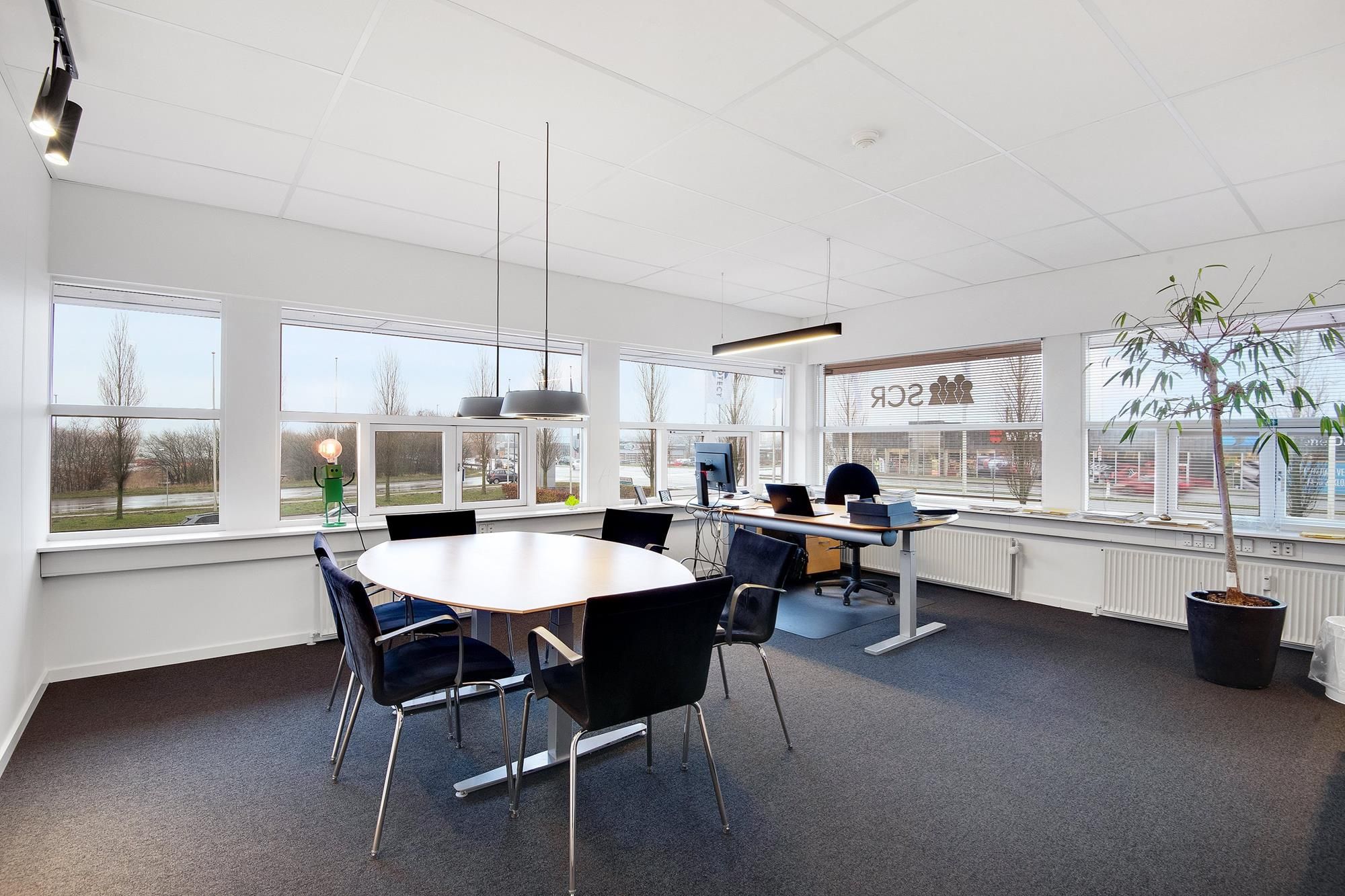 Kontorlokaler med lyse vinduer, moderne møbler og en plante ved siden af vinduet. Væggene er hvide, og der er en hvid