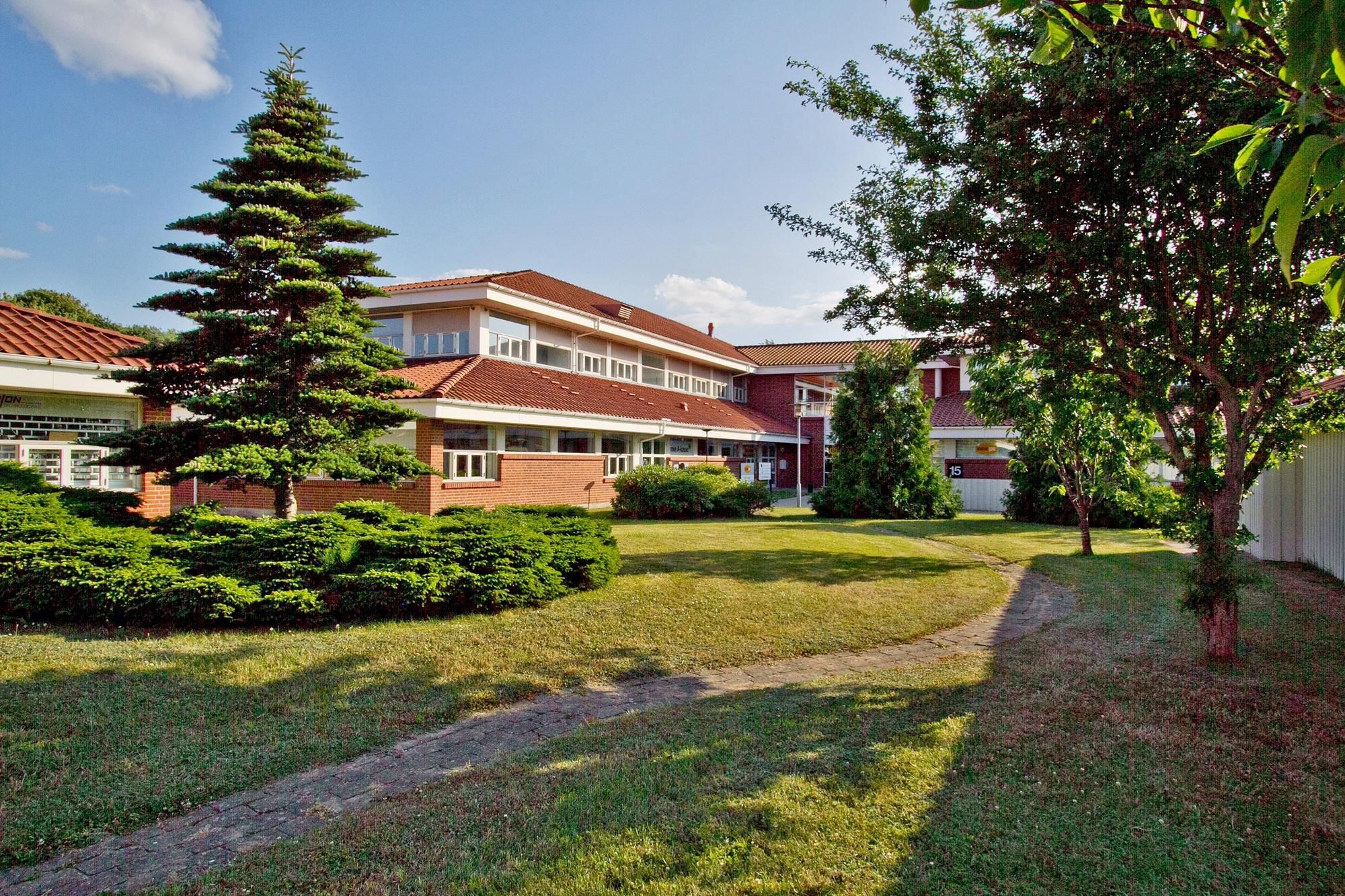 En stor bygning med mange vinduer og røde tagfliser, lyse kontorlokaler, grønne træer og buske, og en sti gennem en grøn