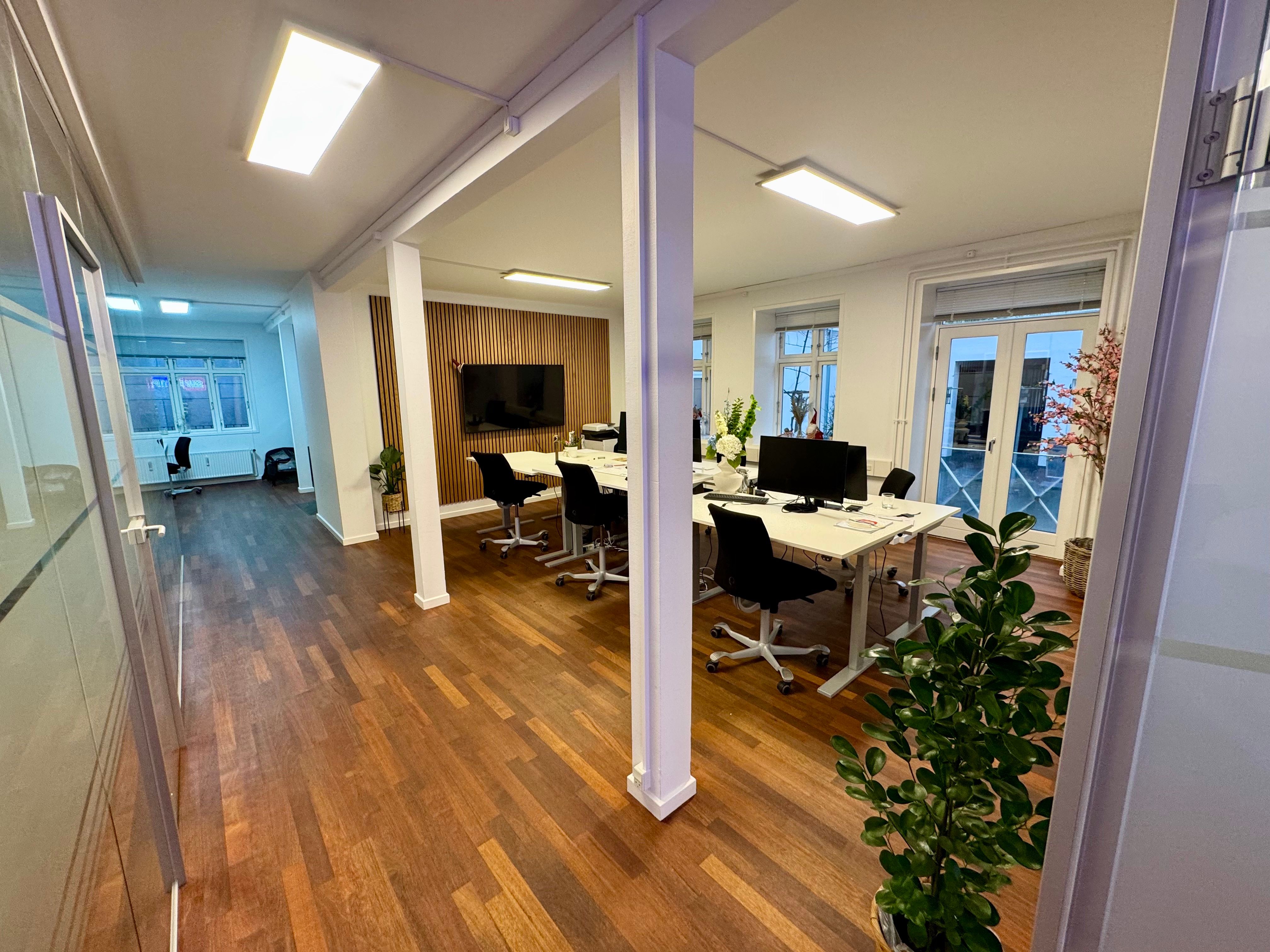 Lukket kontor med lyse trægulve og hvide vægge, moderne møbler og plante ved vinduet, og en lille tv-skærm i enden af rummet