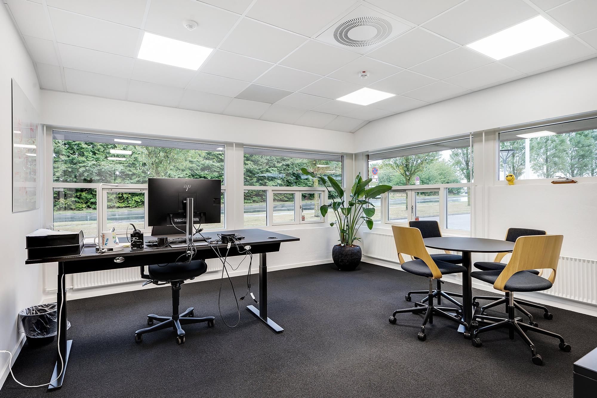 Inde i et kontor med moderne møbler og skrivebord, to små runde borde med fire stole, skrivebord med computer, en plante og