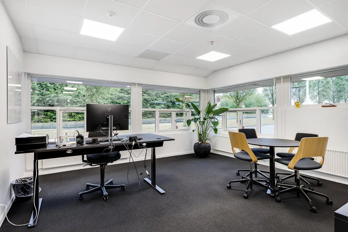 Inde i et kontor med moderne møbler og skrivebord, to små runde borde med fire stole, skrivebord med computer, en plante og