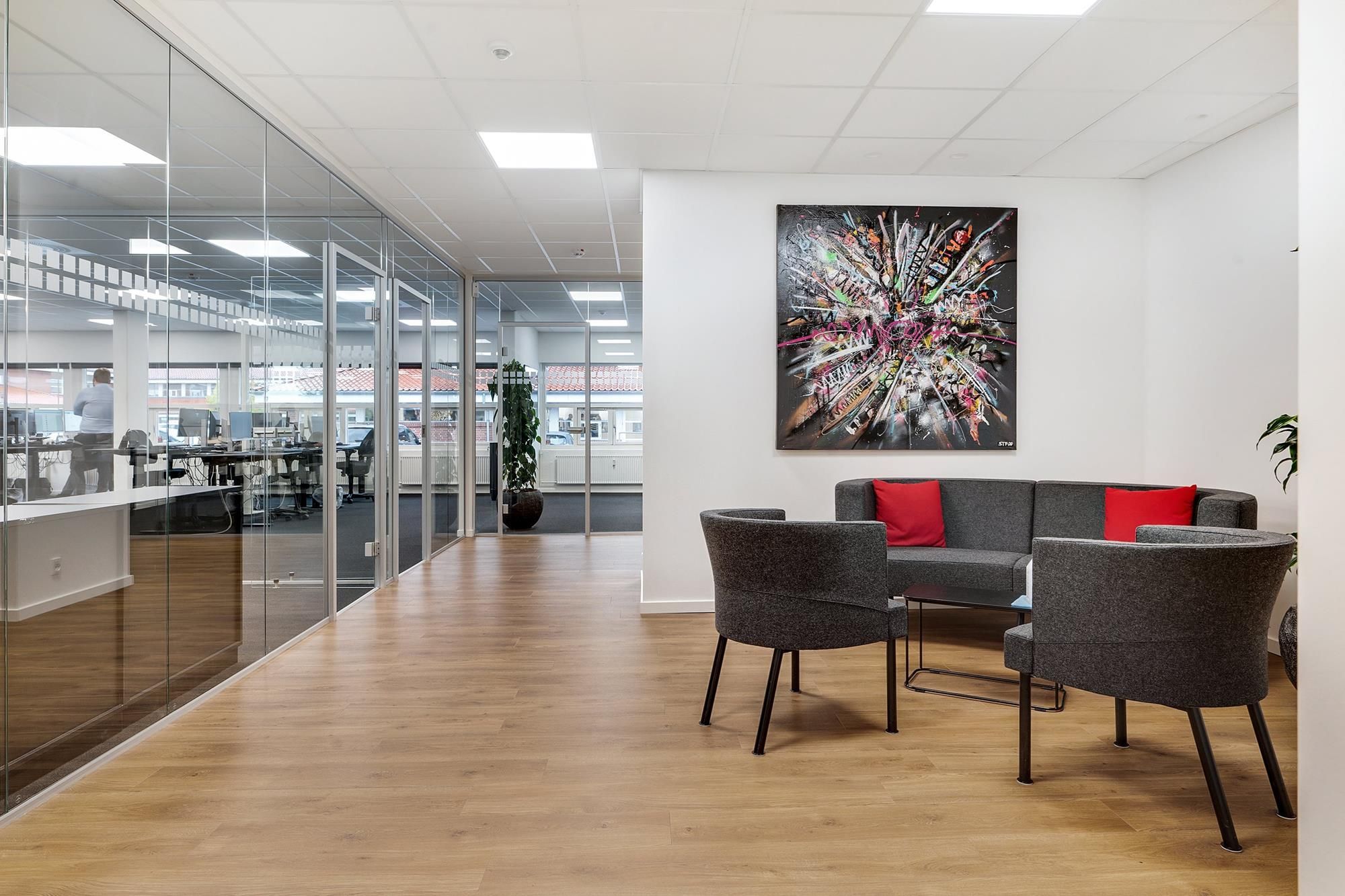 Modernt kontor med trægulv og glasvægge, sofa med røde puder og kunstværk på væggen