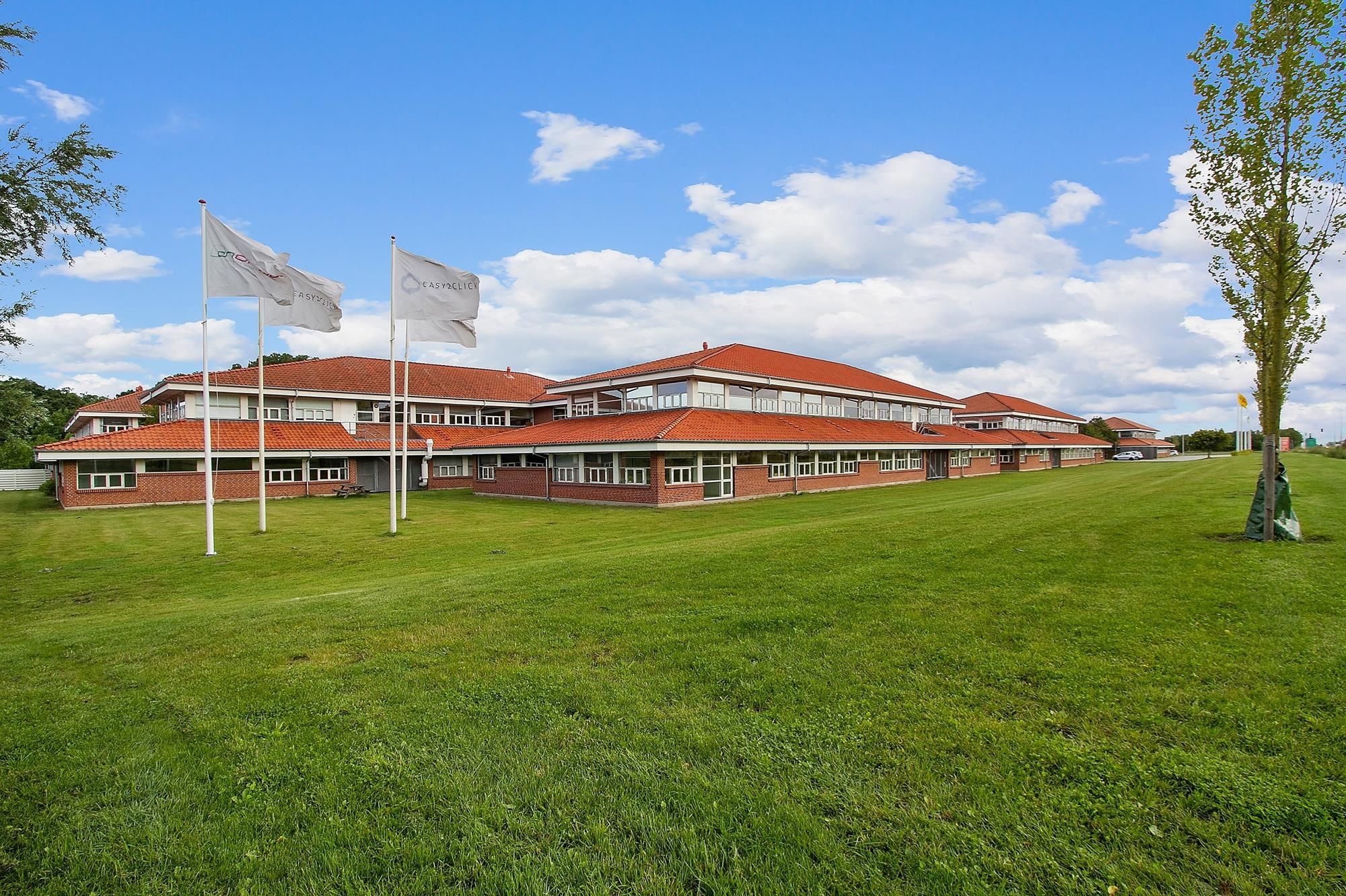 Forretningsbygning med røde tag og store vinduer, omgivet af grønne områder, to hvide flag med logo og en bilparkeringsplads