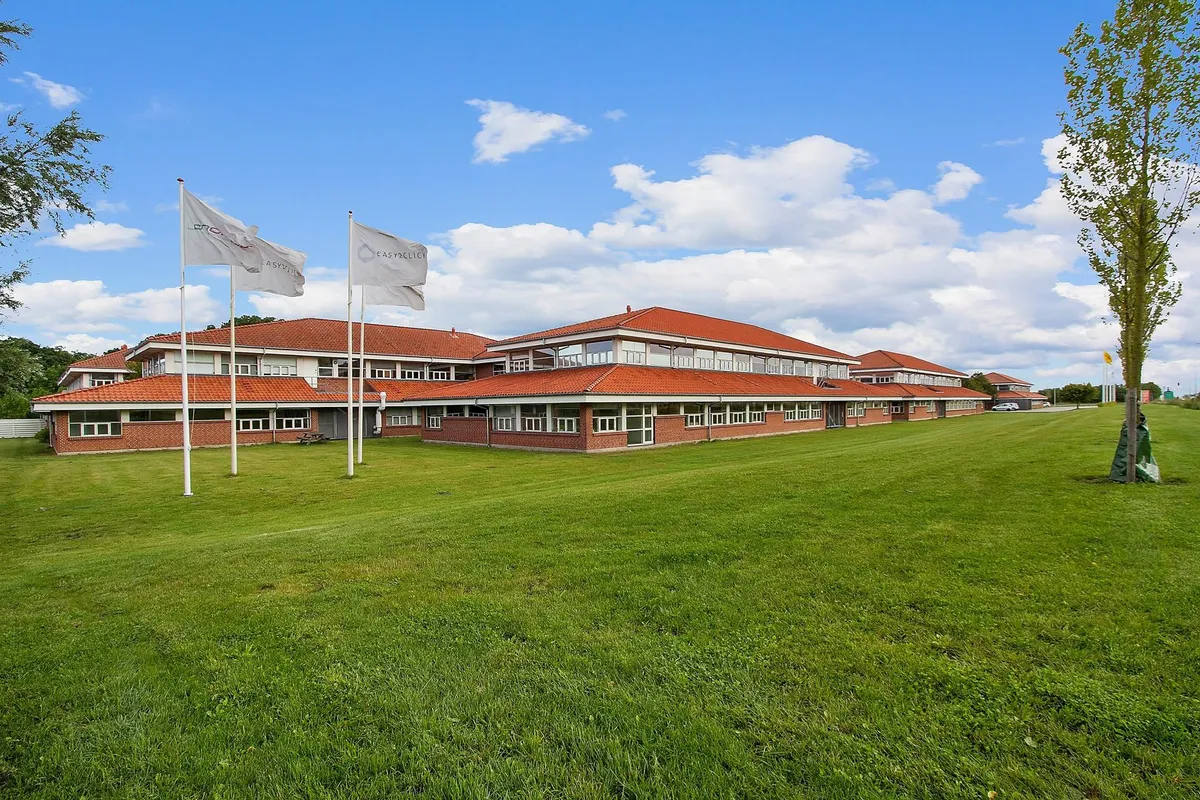 Forretningsbygning med røde tag og store vinduer, omgivet af grønne områder, to hvide flag med logo og en bilparkeringsplads